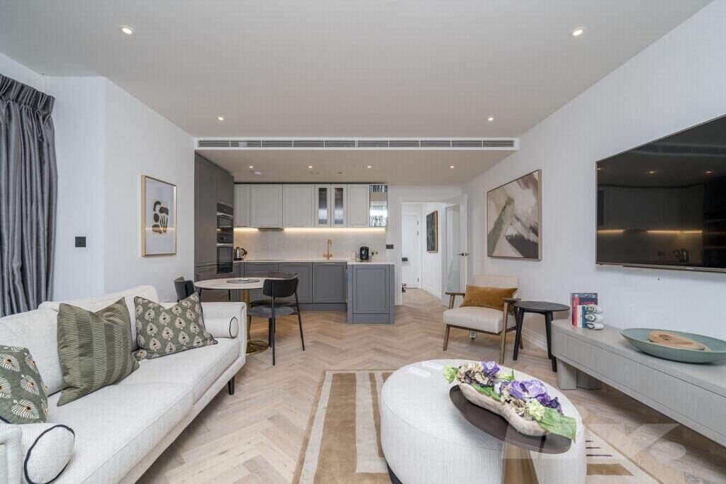 A modern living room with a sofa, a coffee table, and a TV. There is a kitchen in the background with cabinets and a countertop.