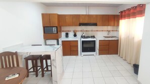A compact kitchen with wooden cabinets, a microwave, a stove, and a sink. There is a dining table with chairs and a remote control on it. - Charming 1-bedroom apartment in lovely Santa Cruz with WiFi, AC (Santa Cruz de la Sierra)