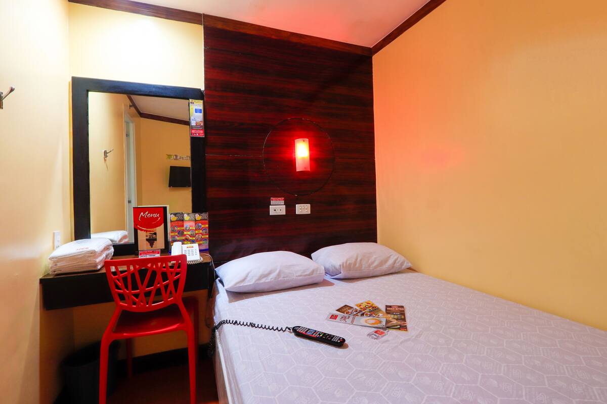 Une chambre d’hôtel comprenant un lit, un bureau avec une chaise, un miroir et un luminaire fixé au mur.