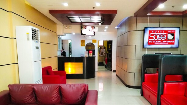 Un vestíbulo de hotel con mostrador de recepción, zona de estar con asientos rojos y un televisor mostrando un anuncio del hotel.