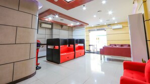 Un hall de hotel con diseño moderno, que presenta una zona de estar en rojo y negro, un mostrador de recepción y un techo luminoso con tragaluces.
