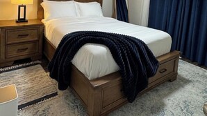 Habitación de hotel con cama de madera, mesita de noche con lámpara, alfombra y ventana con cortinas.