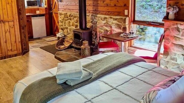 L’intérieur d’un chalet accueillant, avec une cheminée en pierre, un lit recouvert d’une courtepointe, un plafond en bois et une vue sur l’extérieur par une fenêtre.