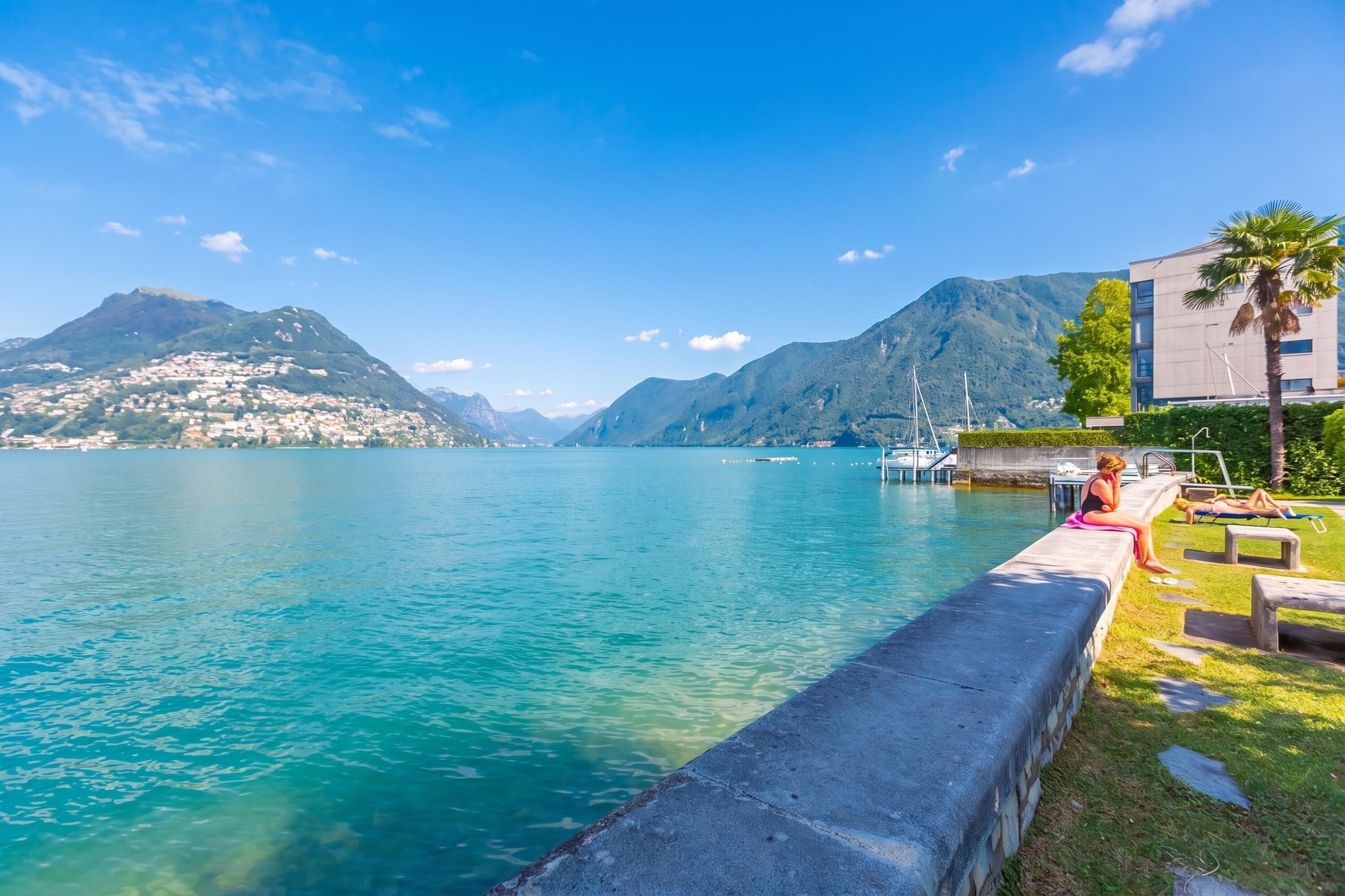 A waterfront view with a clear lake, mountains in the background, and a modern building with a swimming area.