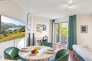 A modern hotel room with a dining area, a bed, a large window with a view of the outdoors, and a flat-screen TV mounted on the wall.