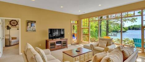 A spacious living room with a large window offering a view of a lake and greenery. There is a television on a wooden stand, and a sofa set is arranged facing the window.