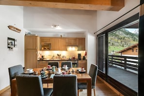 Een eetruimte met een houten tafel voor vier personen, een keuken op de achtergrond en een groot raam met uitzicht op de bergen.