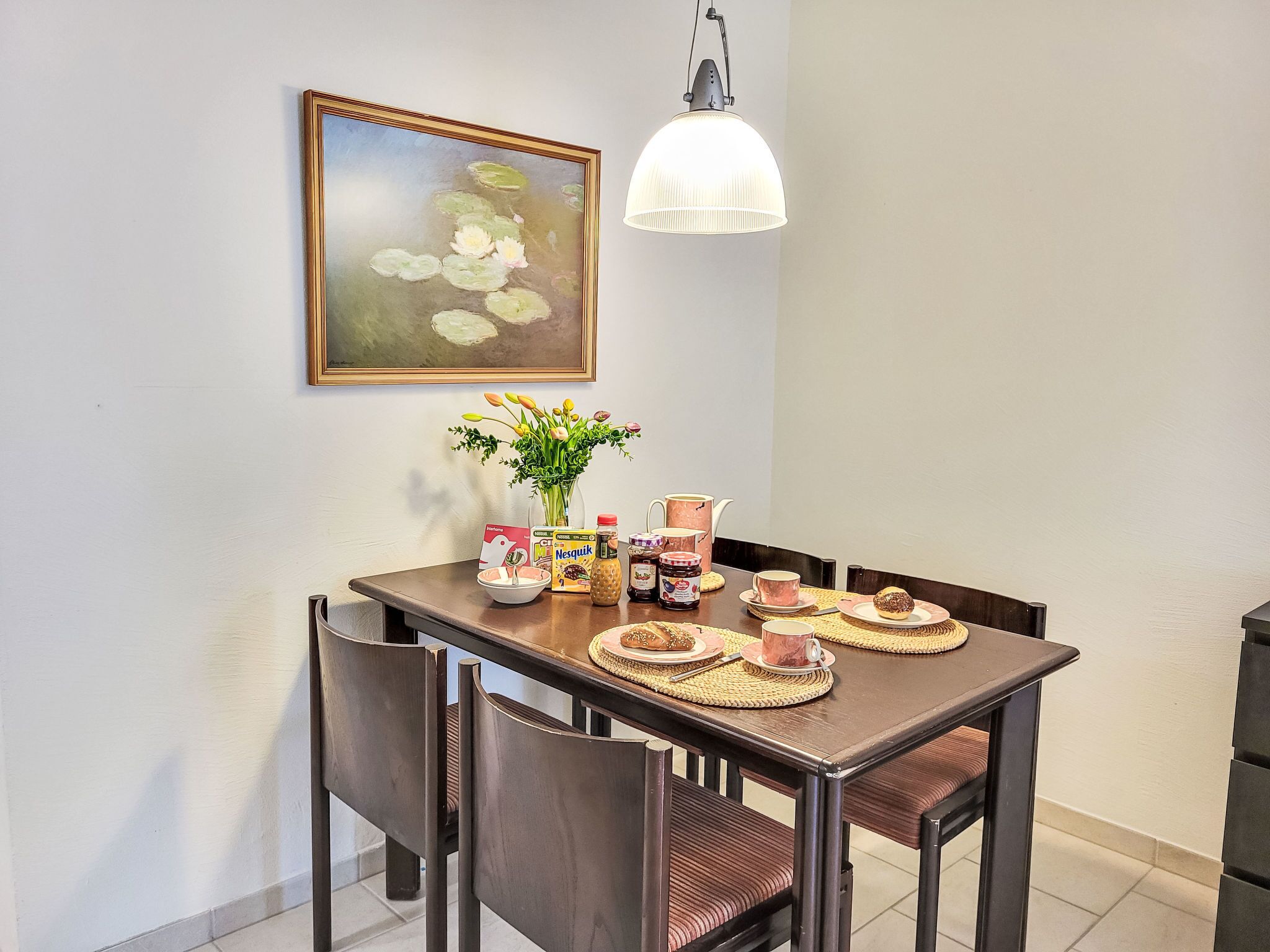 Un'area pranzo con un tavolo apparecchiato per due persone, un vaso con fiori e un quadro appeso al muro.
