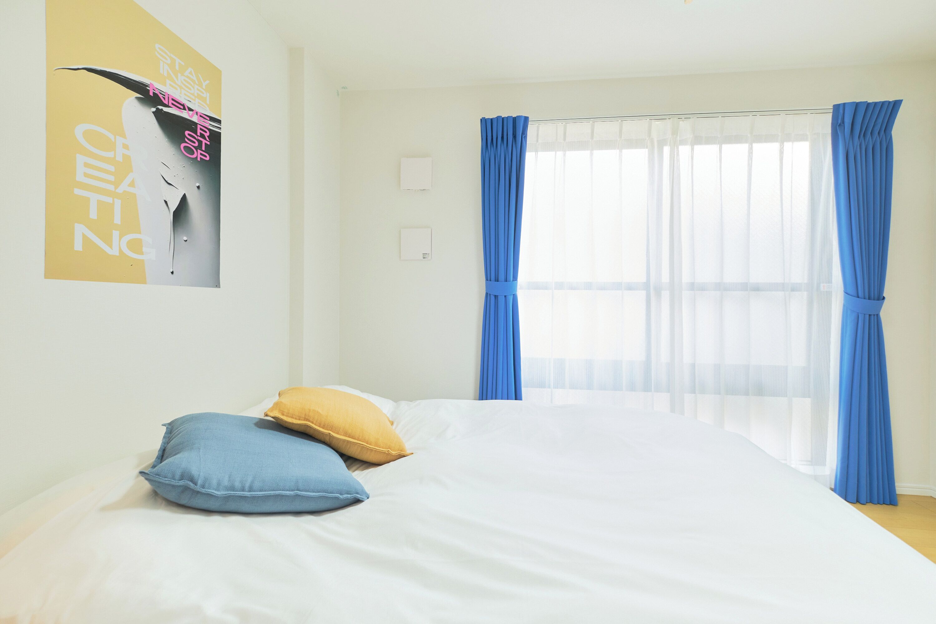 Uma cama bem arrumada, com almofadas azuis e amarelas, uma parede branca e uma janela com cortinas azuis.
