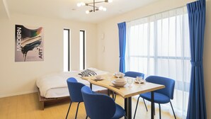 Une pièce comprenant un lit, une table à manger avec des chaises, un luminaire au plafond et une grande fenêtre avec des rideaux.