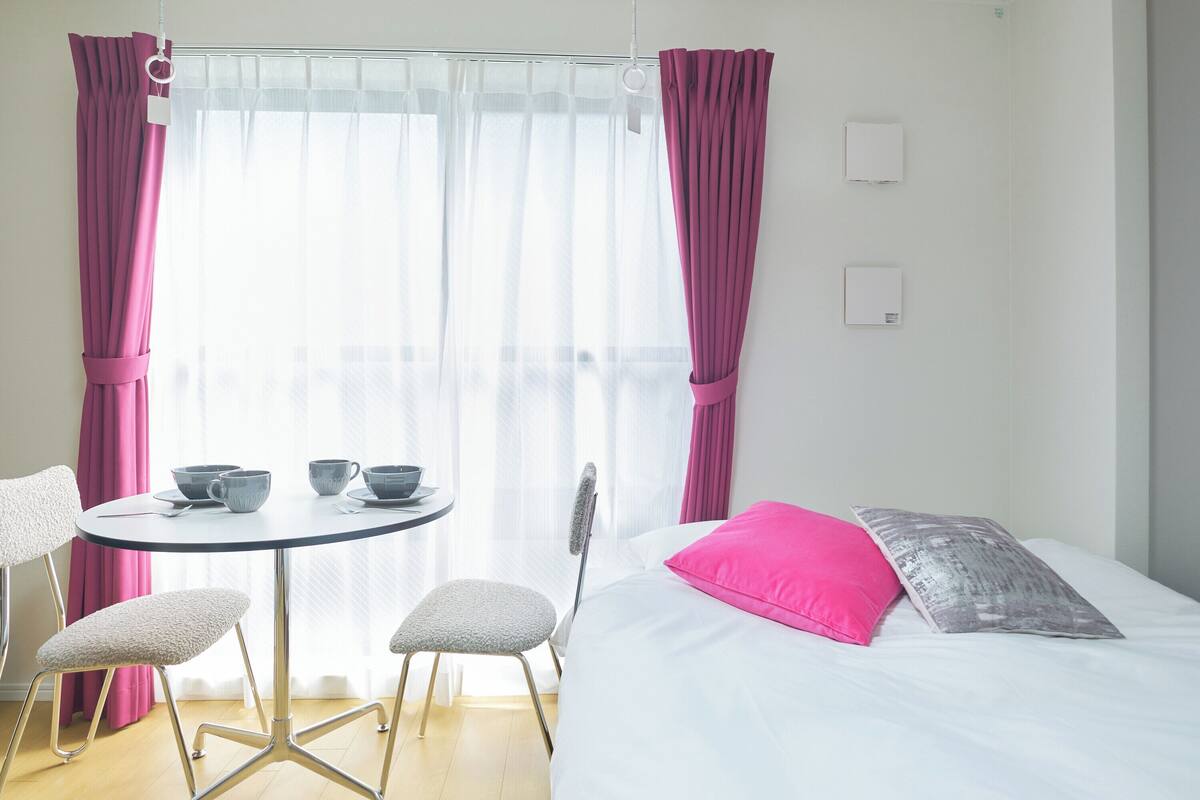 Una habitación con una cama, una mesa redonda con sillas, un ventanal con cortinas translúcidas y un interruptor de luz en la pared.