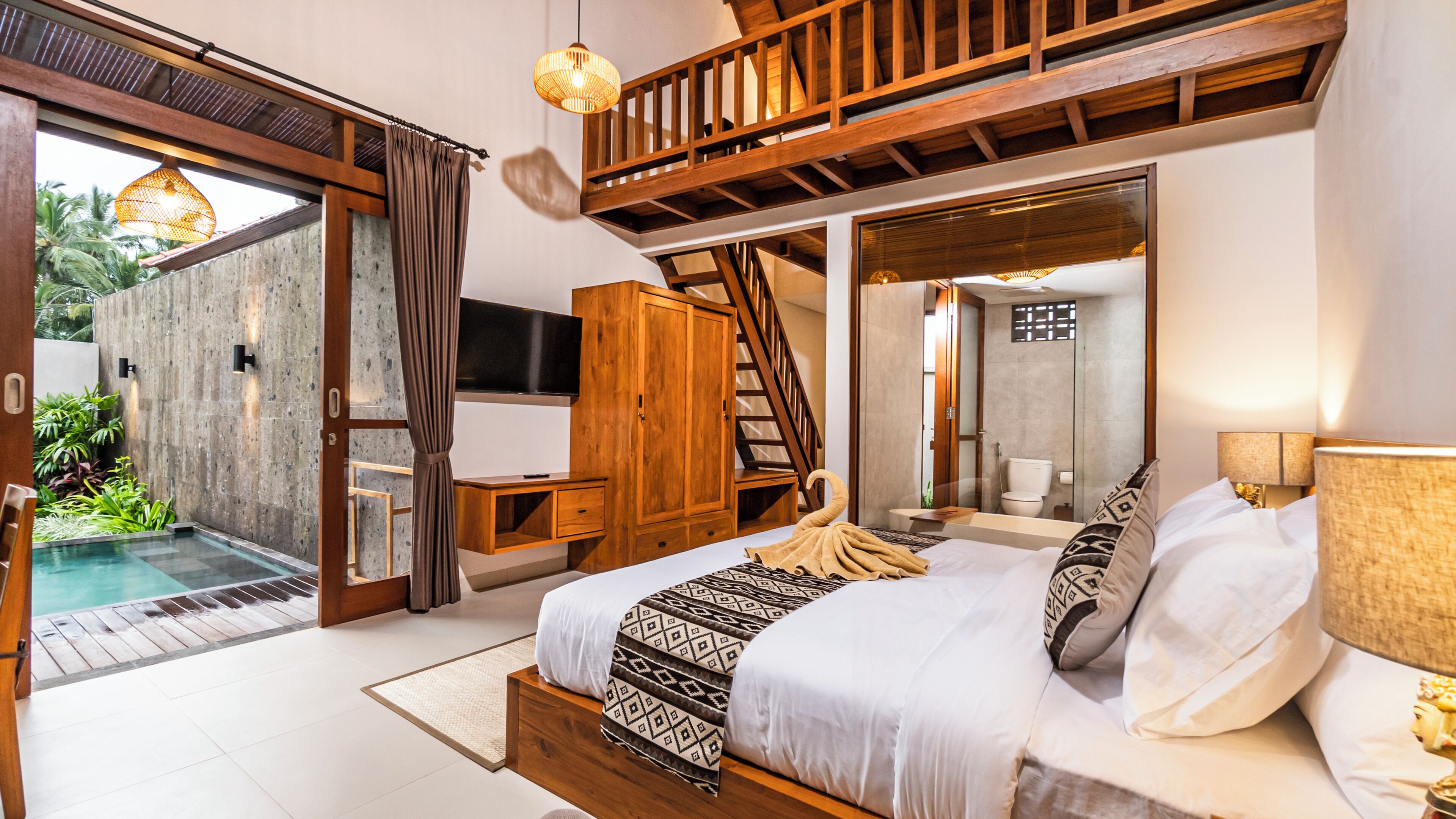 Quarto com uma cama grande, televisão, guarda-roupa de madeira e banheiro visível por uma porta aberta.