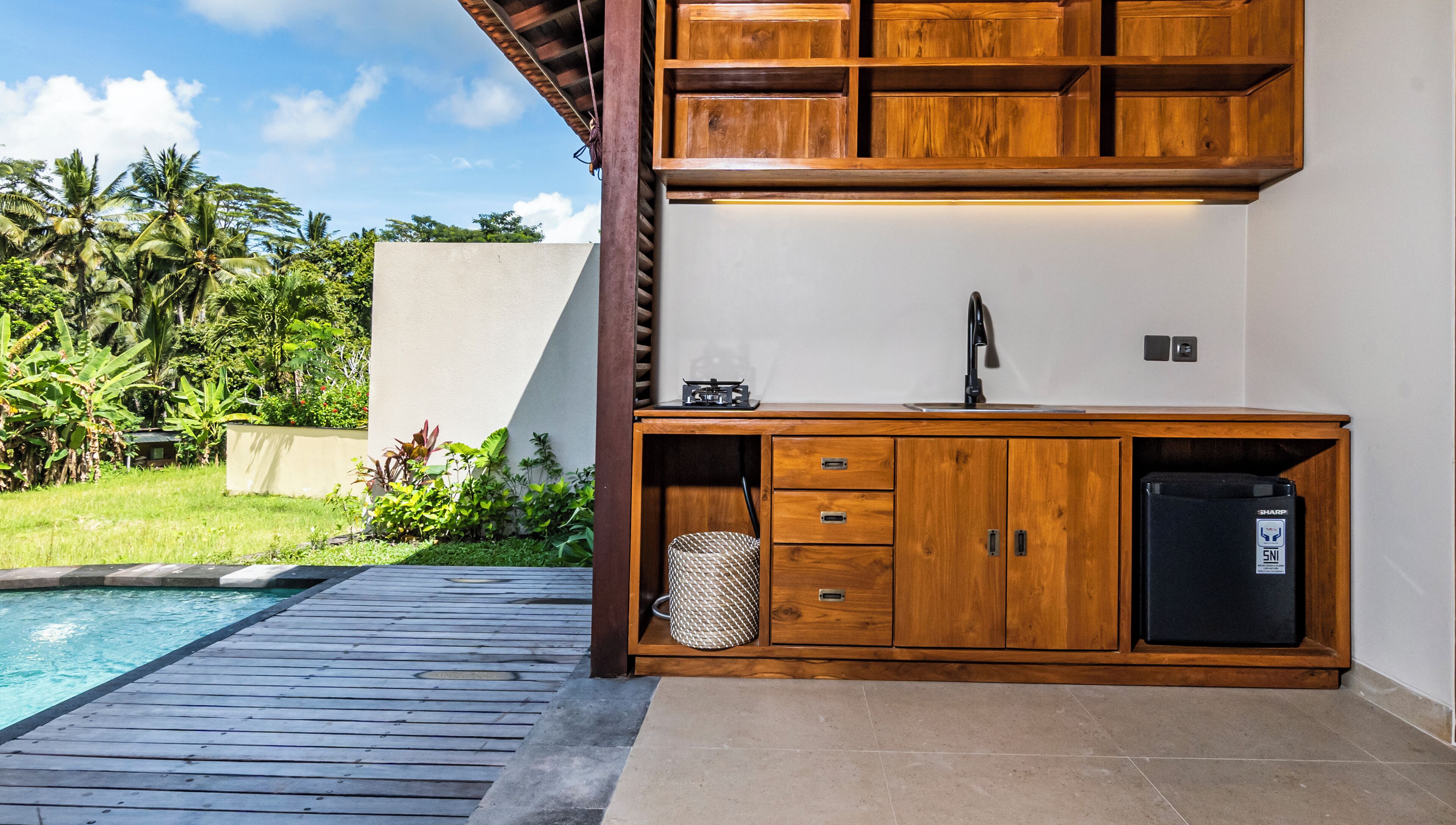 A wooden outdoor kitchen with a built-in sink and a refrigerator, adjacent to a swimming pool and surrounded by lush greenery.