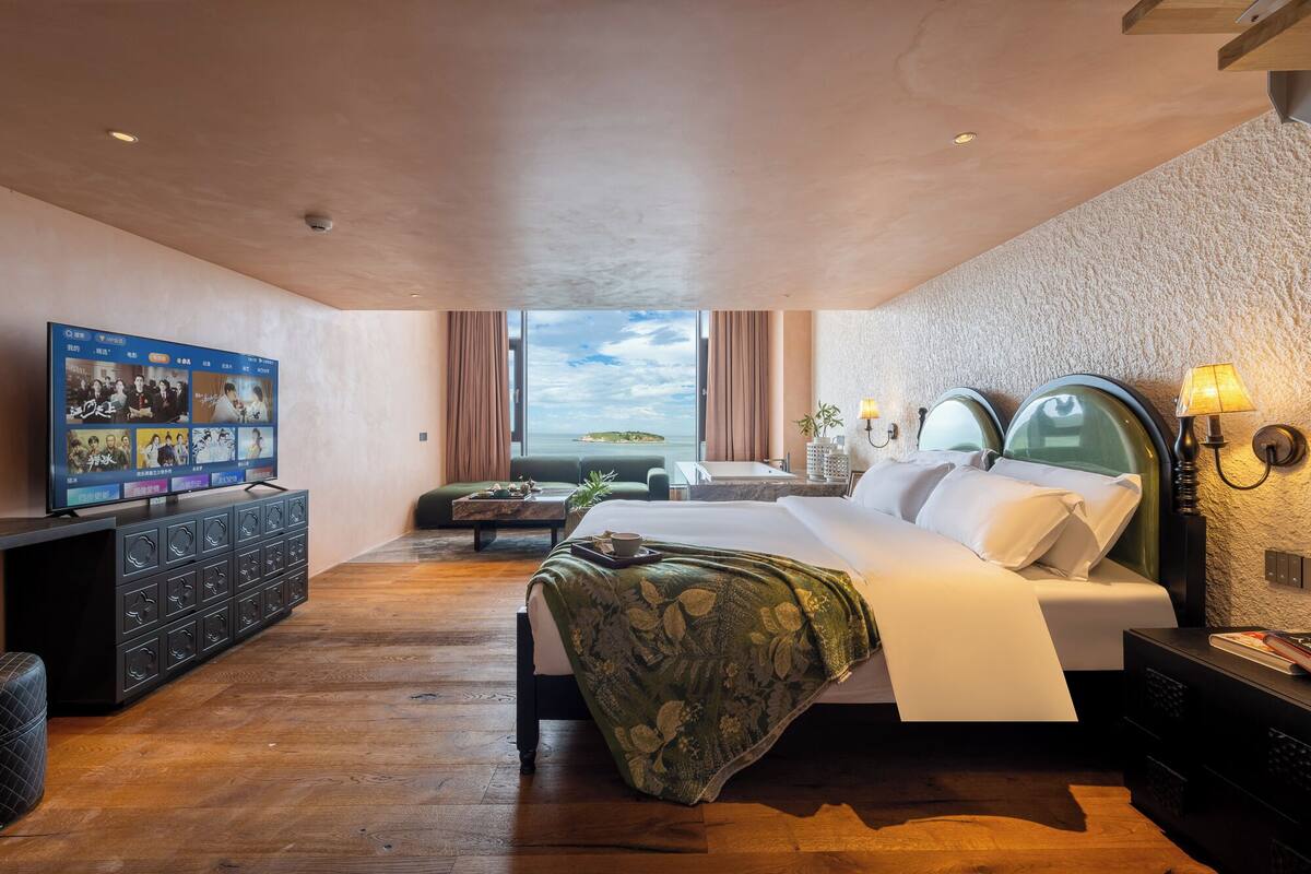 A modern hotel room with a large bed, a television, a desk, and a view of the sea.