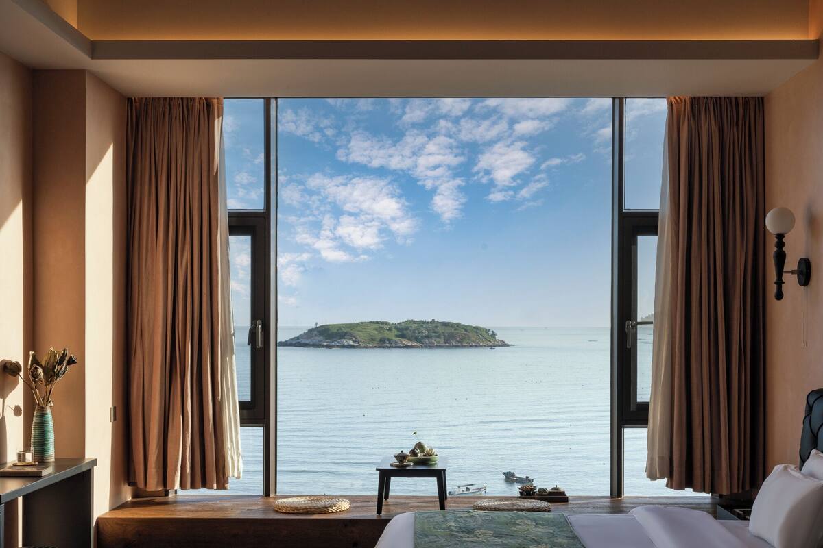 A bedroom with a large window overlooking a calm sea and a small island.
