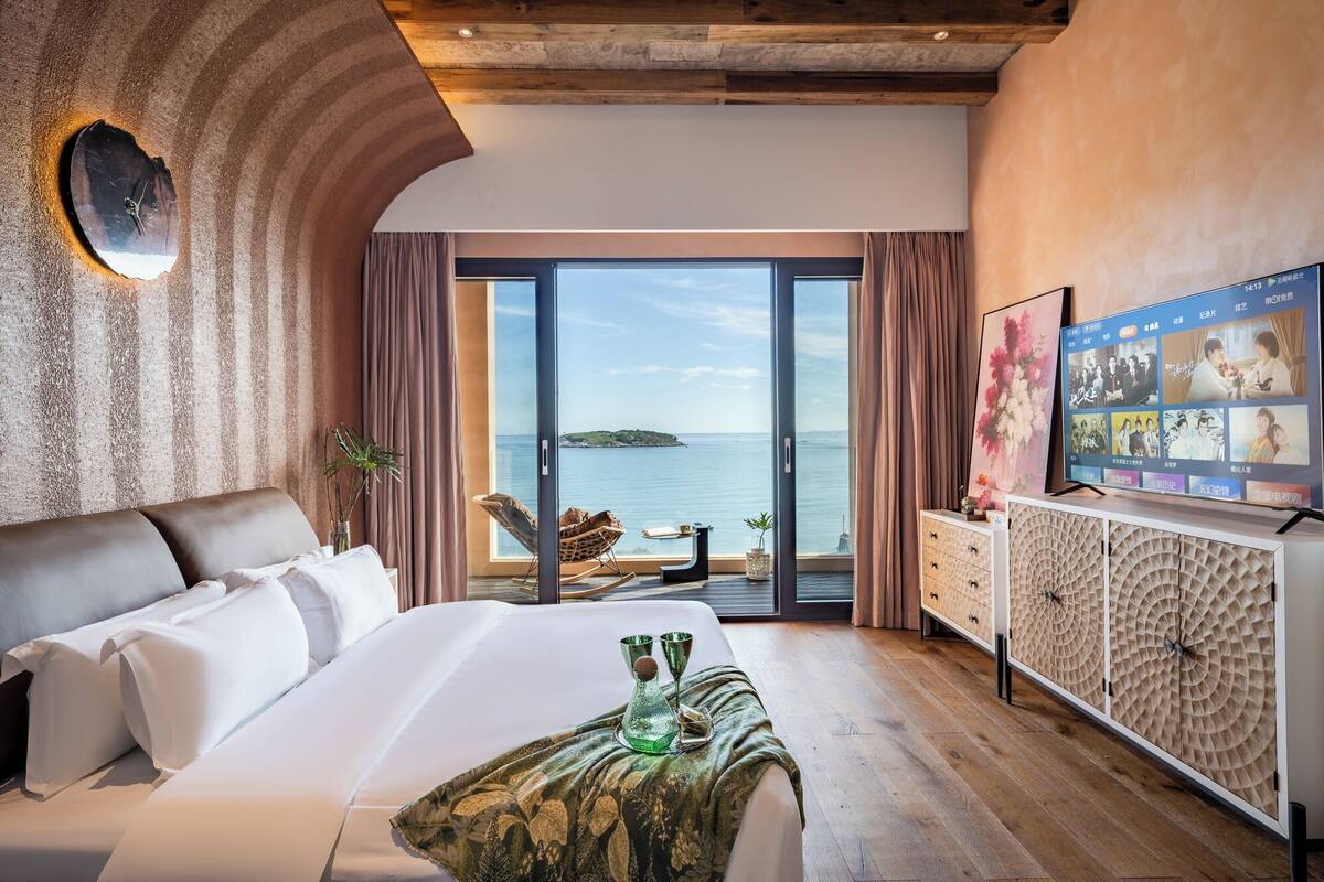 A hotel room with a large bed, a view of the sea, a television, and a wooden ceiling.
