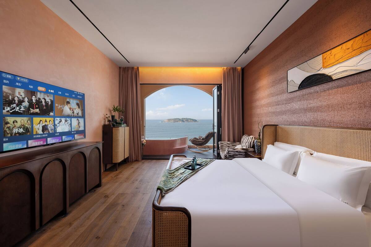 A hotel room with a large bed, a television displaying a program, a view of the sea, and a wooden wall.