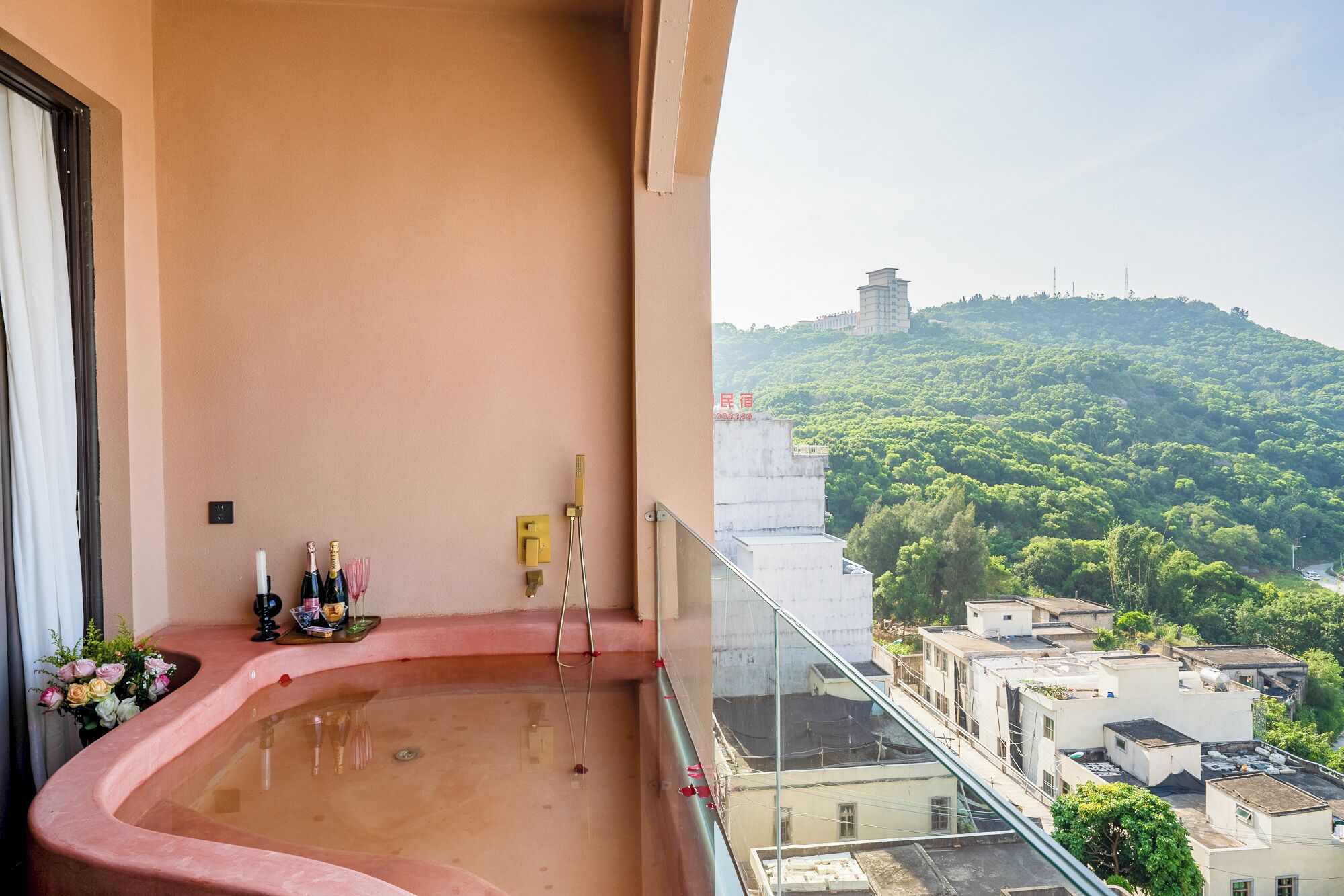 A balcony with a hot tub, a glass railing, and a view of a green hillside with buildings.