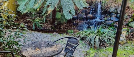 Een terras met een ronde tafel en twee stoelen, omgeven door weelderige begroeiing en een kleine waterval.