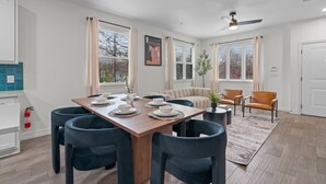 Un coin repas avec une table en bois dressée pour quatre personnes, entourée de chaises bleu foncé. On aperçoit un canapé beige et une chaise orange à l'arrière-plan.