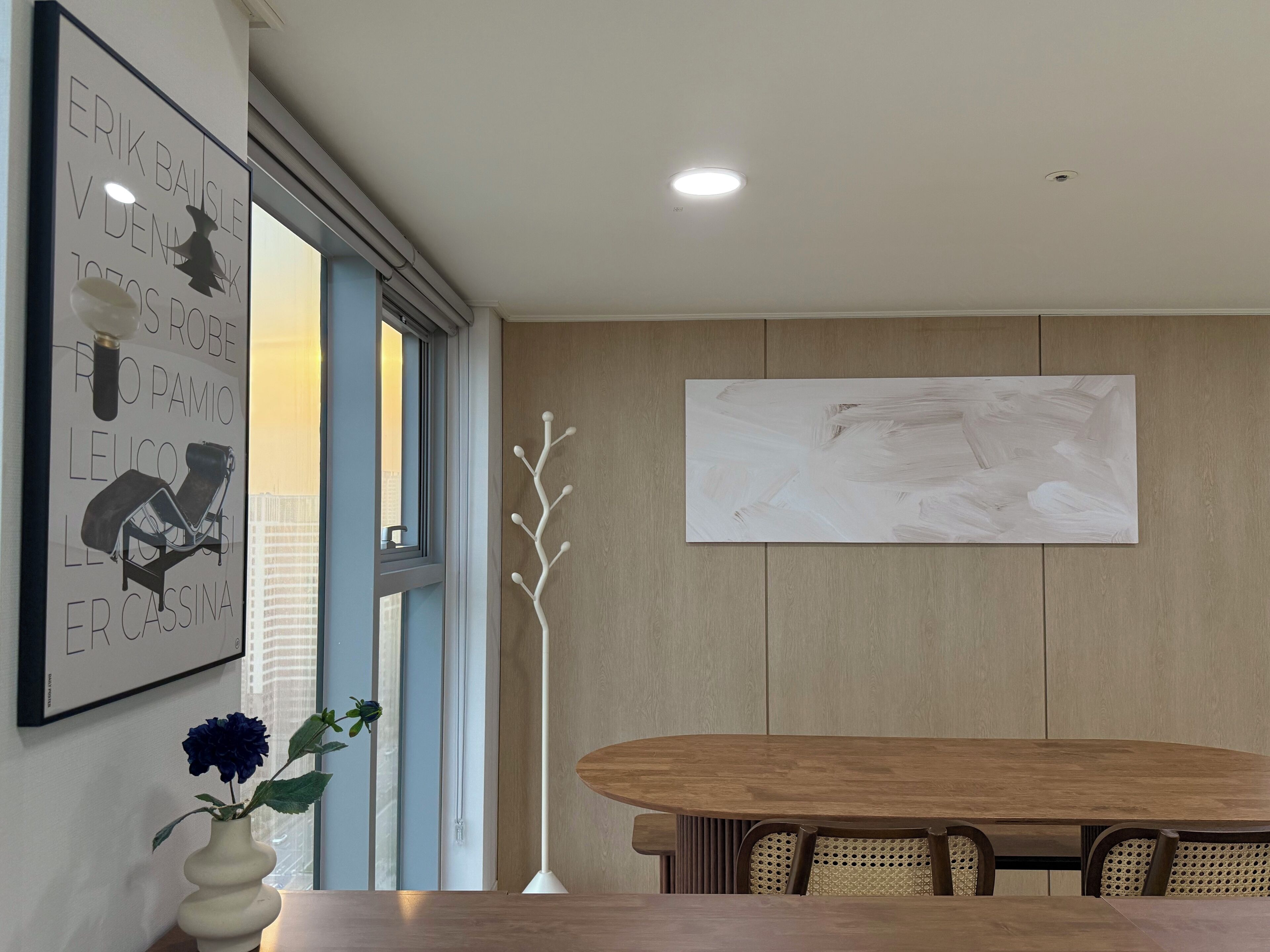 A dining area with a wooden table, a white vase with flowers, a wall-mounted abstract painting, and a framed picture with text and a silhouette.