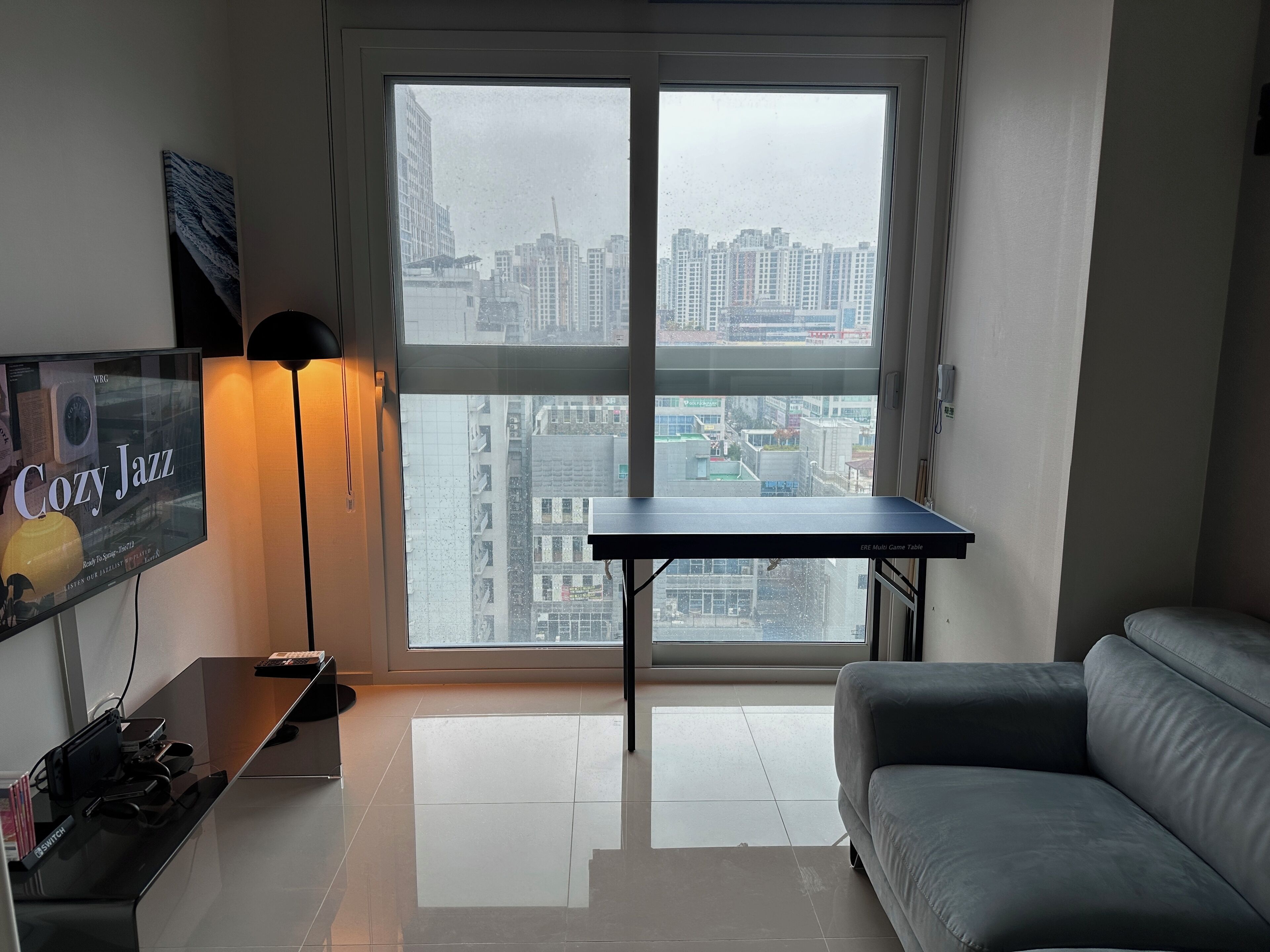 A modern living room with a large window, a grey sofa, a small table, and a TV displaying 'Cozy Jazz'.