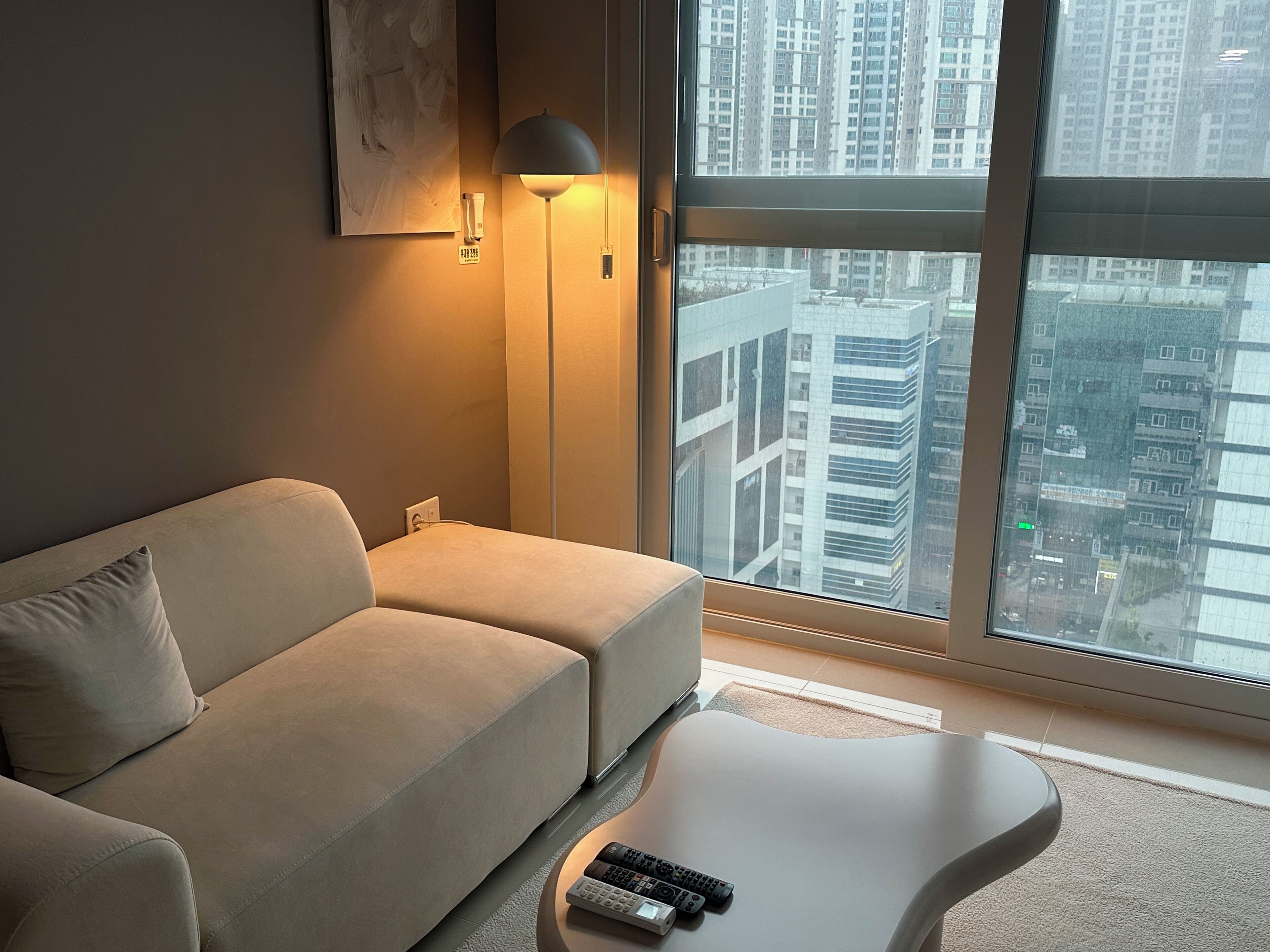 A modern living room with a sofa and a coffee table, a floor lamp, and a large window with a city view.