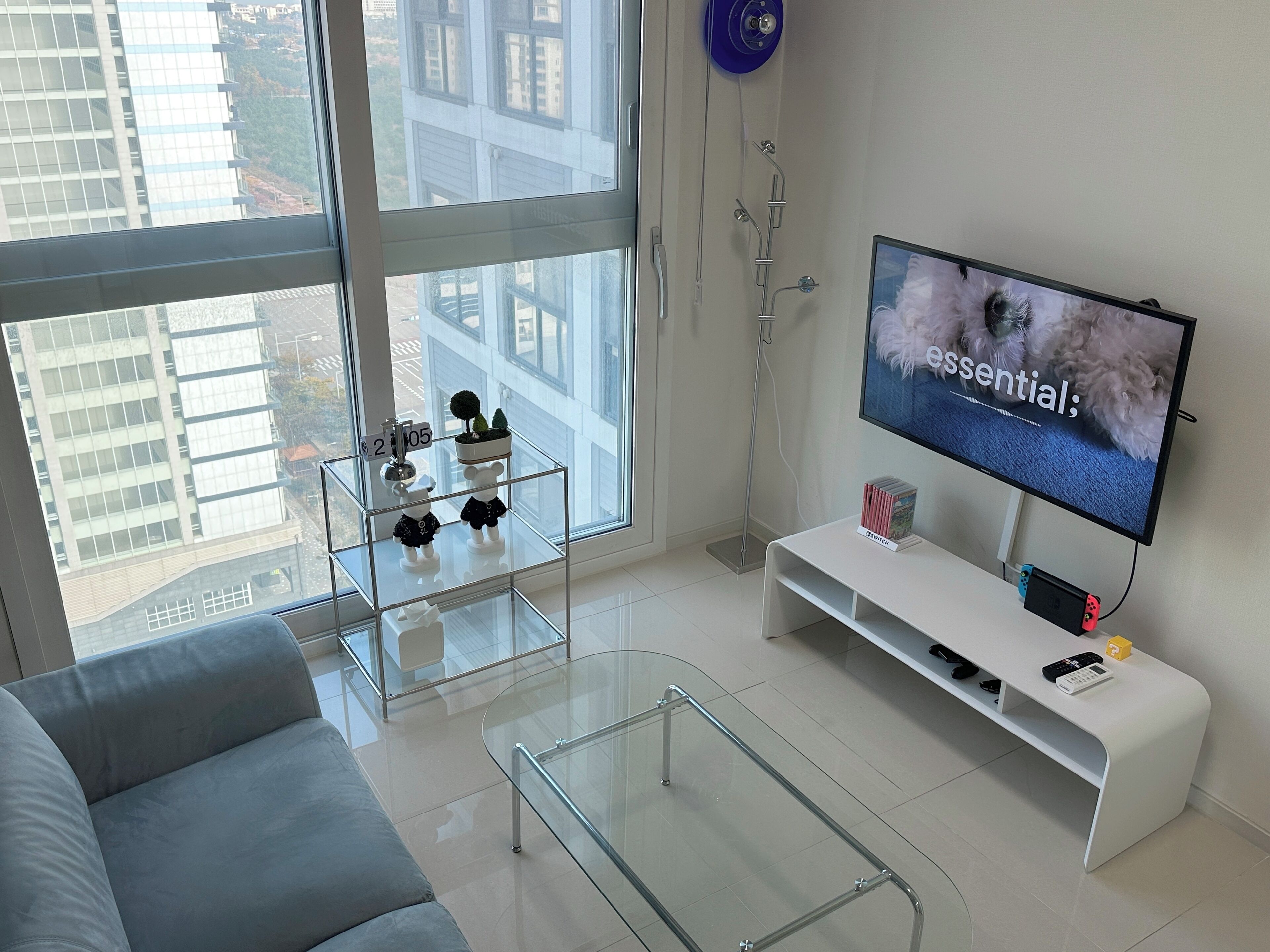 A modern living room with a large window, a glass coffee table, a white sofa, a TV on the wall, and a small shelf with decorative items.