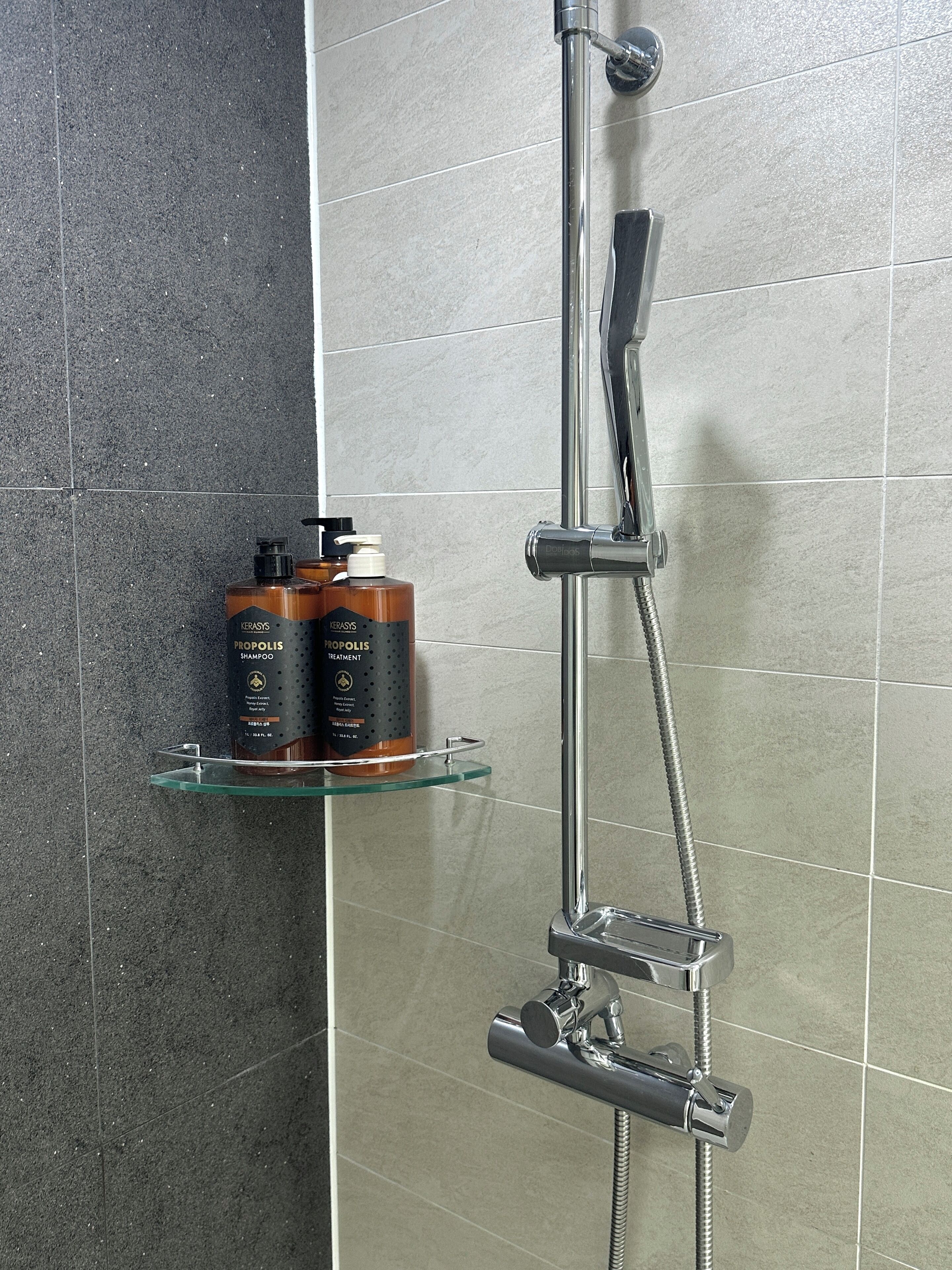 Ducha con cabezal de ducha de mano y grifo empotrado. Hay dos botellas de champú en un estante de vidrio.