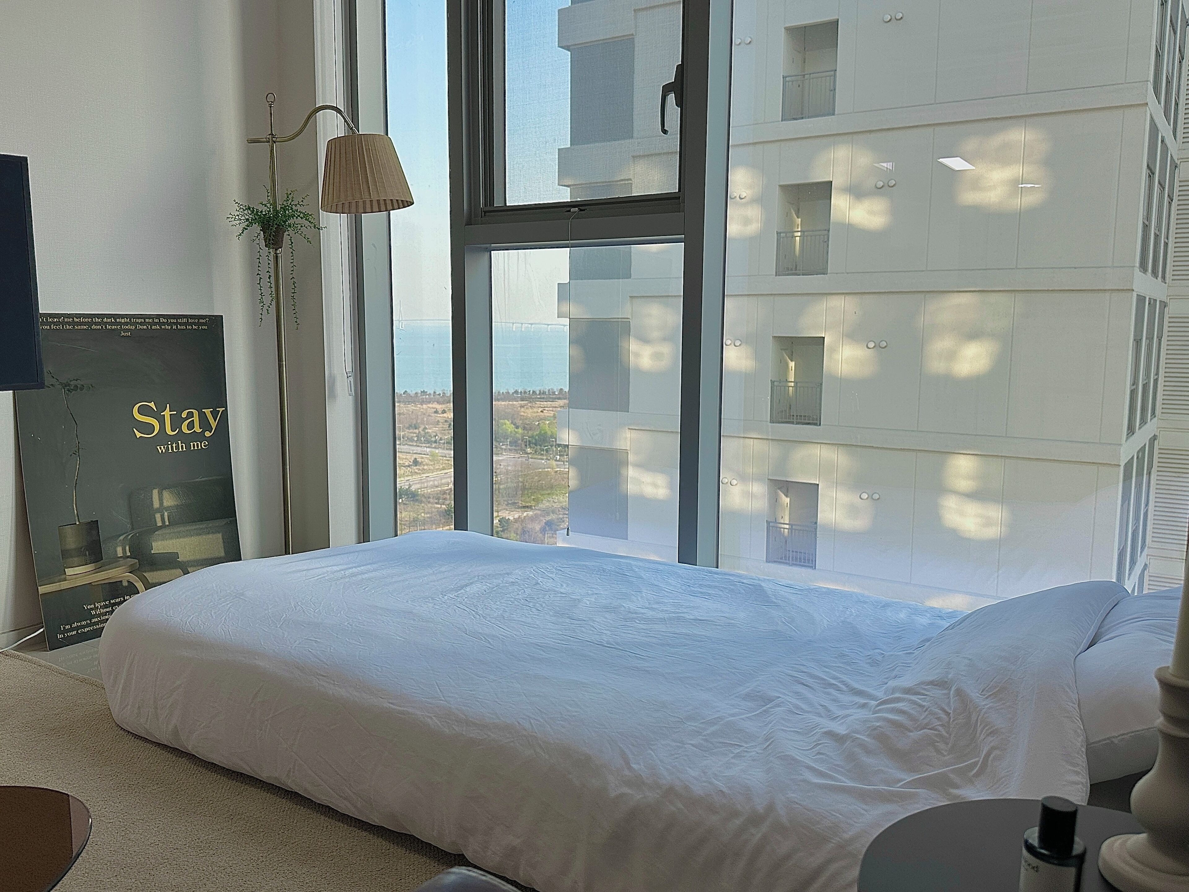 A bedroom with a large window, a bed, a lamp, and a poster on the wall.