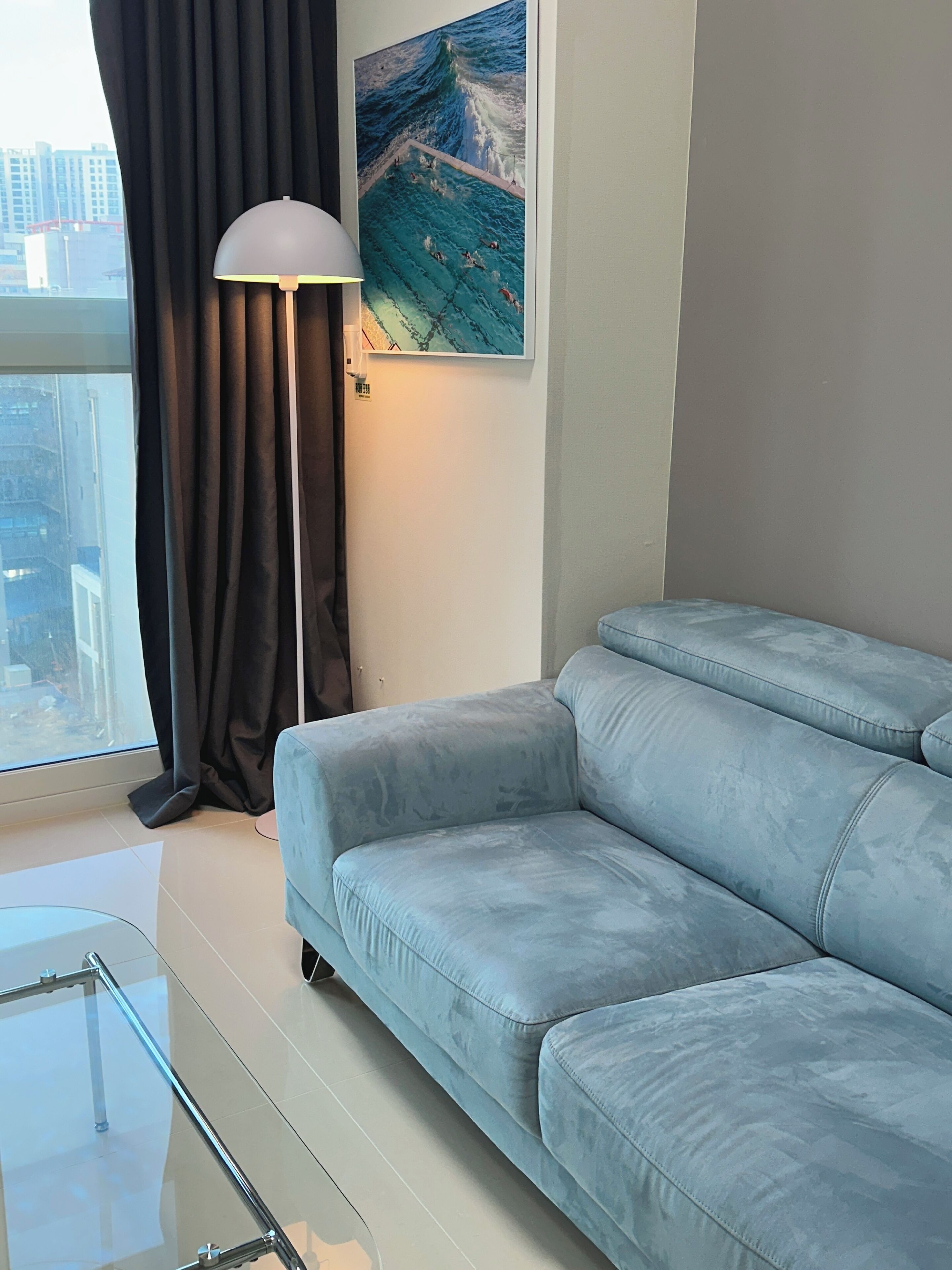 Een moderne woonkamer met een lichtblauwe bank, een vloerlamp en een groot raam met gordijnen.