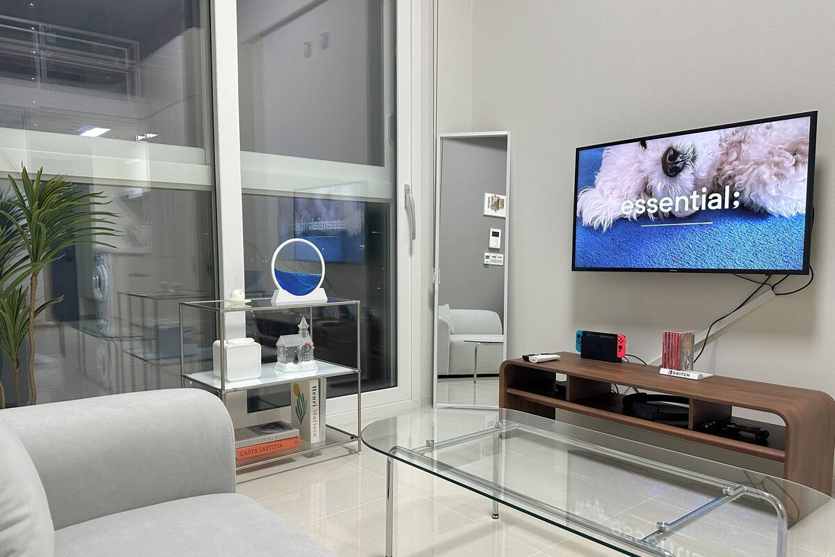 A modern living room with a glass coffee table, a flat-screen TV mounted on the wall, a white sofa, and a wooden entertainment unit.