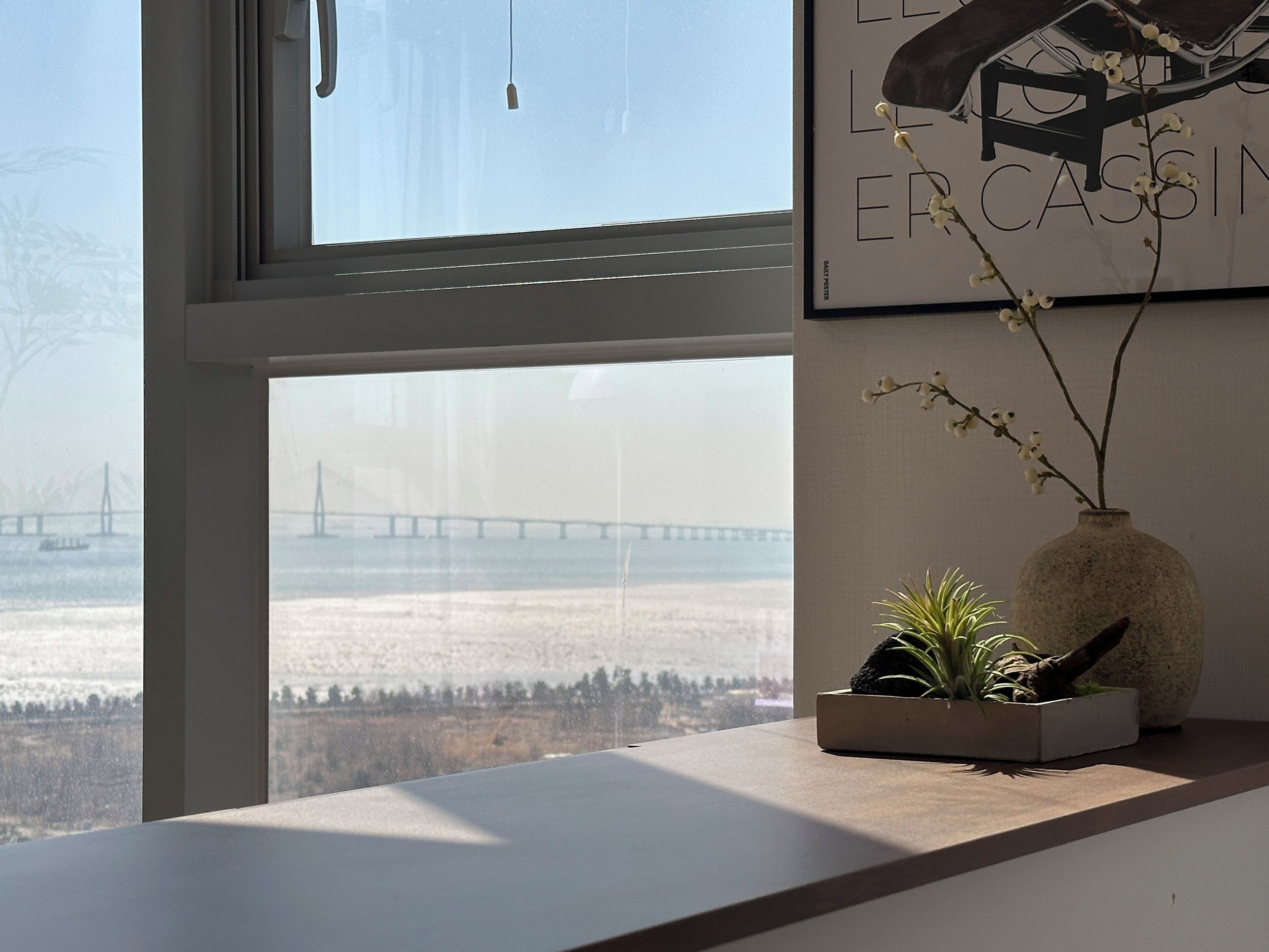 Une pièce dotée d’une grande fenêtre donnant sur un paysage enneigé et un pont. Un tableau encadré orne le mur, et un vase contenant une plante est posé sur le rebord de la fenêtre.