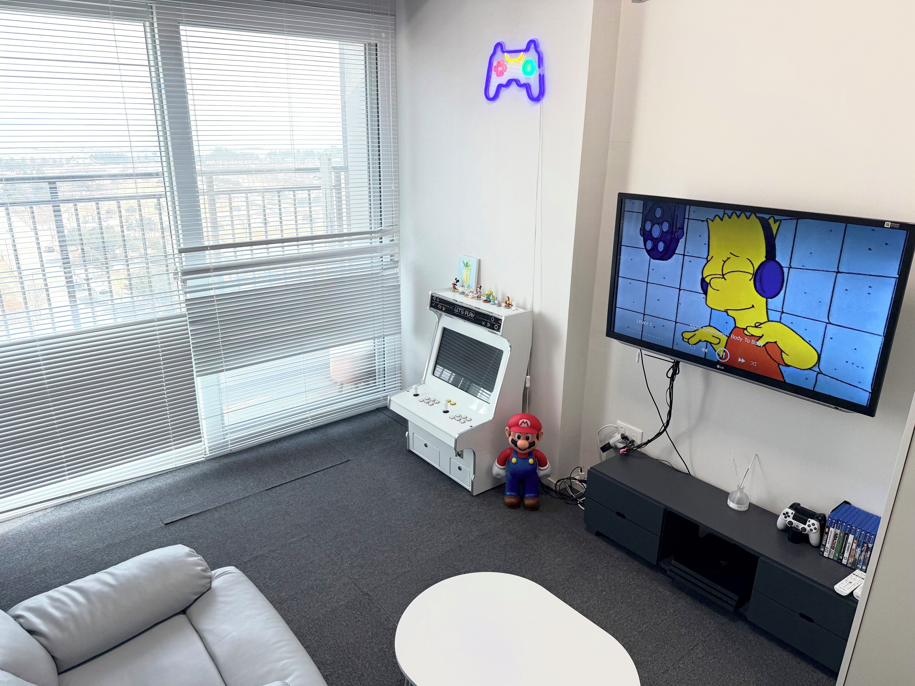 Habitació amb una consola de jocs, un televisor que mostra un personatge de dibuix animat, un peluix i una finestra amb persianes.