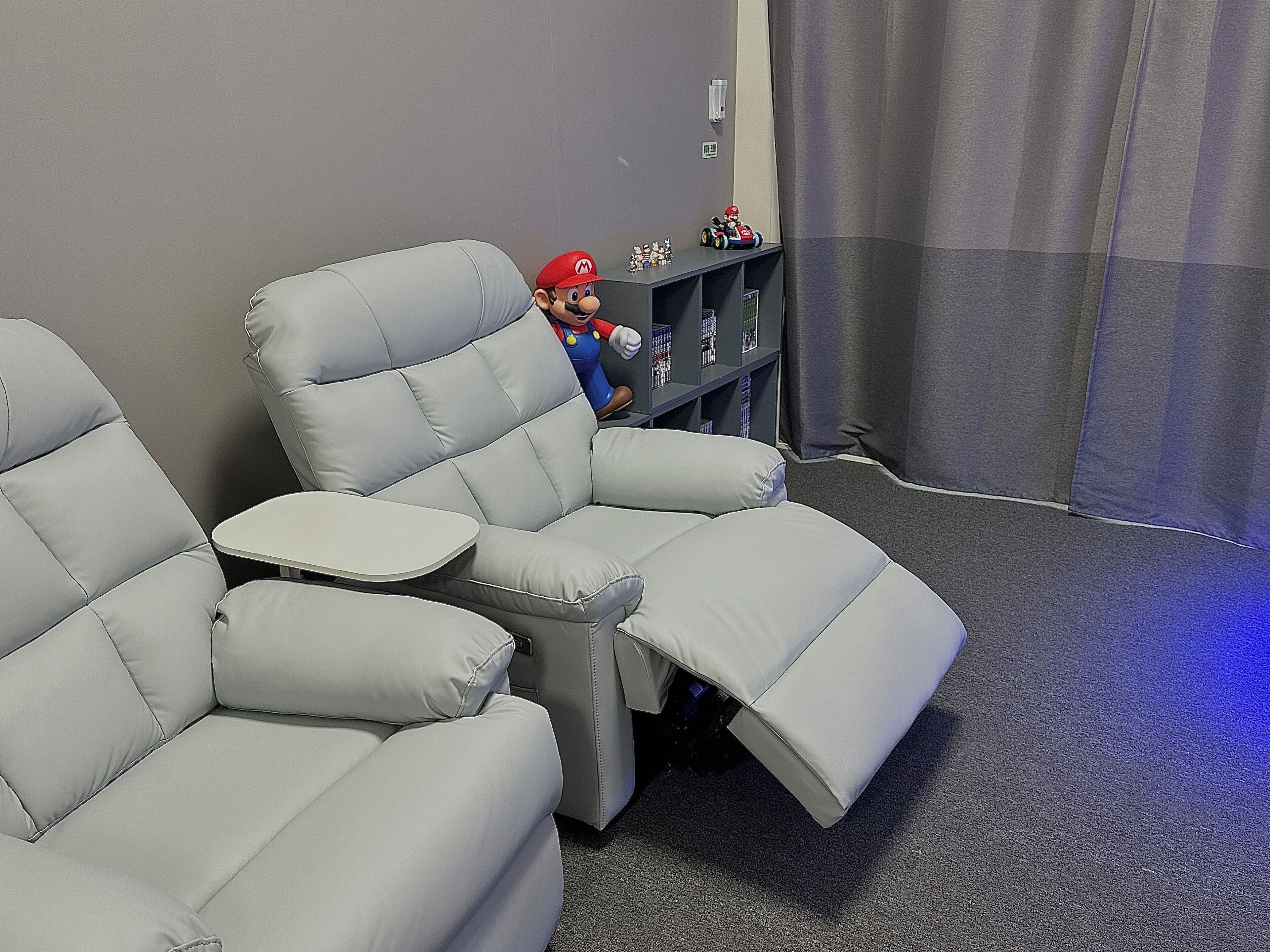 A gray reclining chair with a small table, a shelf with toys, and a gray curtain.