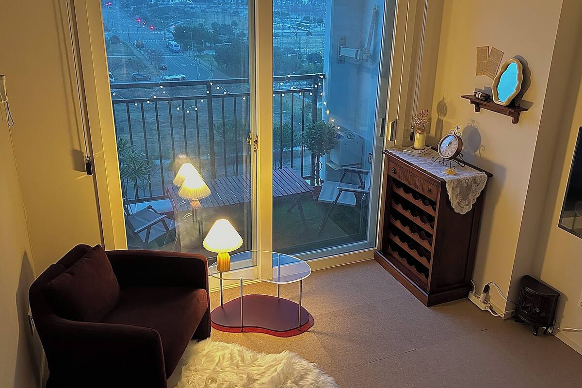 A living room with a sofa, a small table with a lamp, a wooden cabinet with a clock, and a view of a cityscape through a large glass door.