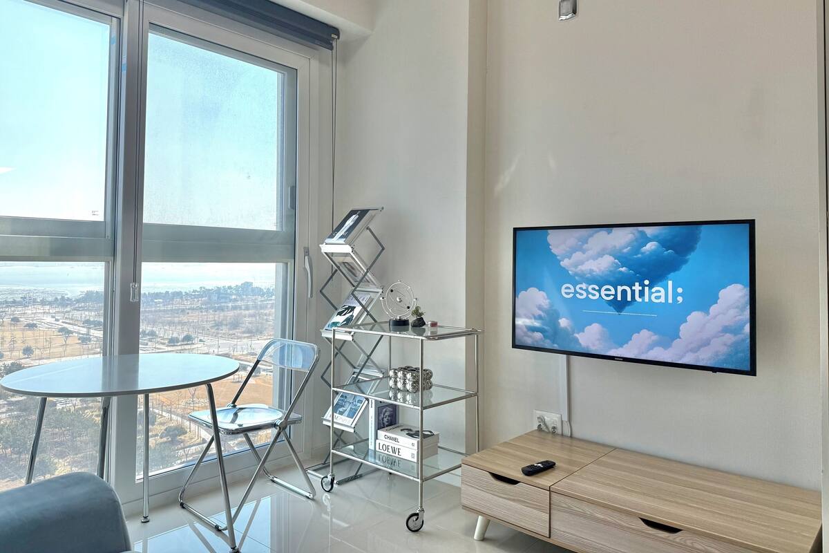 A modern room with a large window, a TV on the wall, a small table, and a glass shelf with books and decorative items.