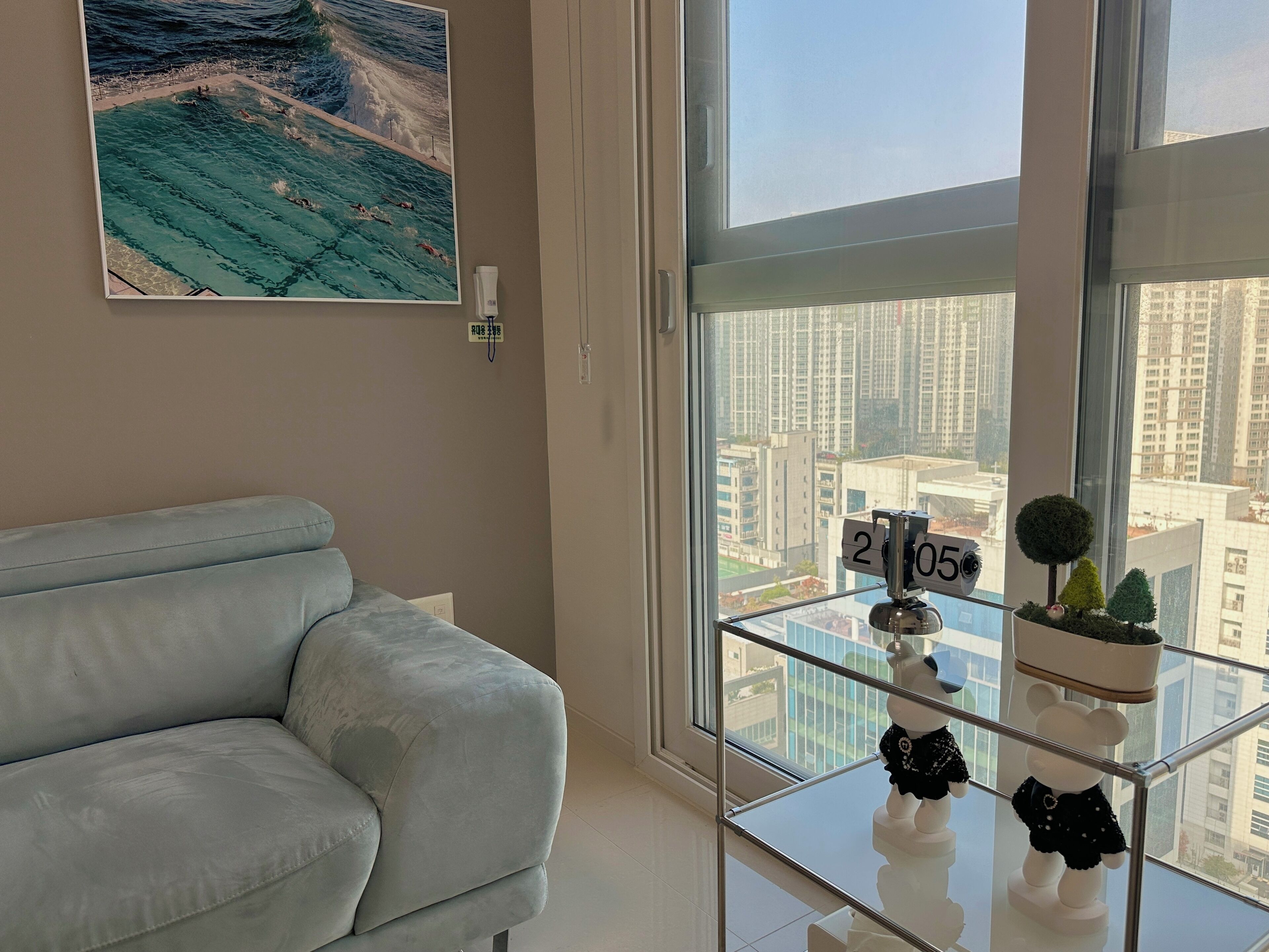 A modern living room with a light grey sofa, a glass coffee table with decorative items, a large window with a city view, and a wall-mounted air purifier.