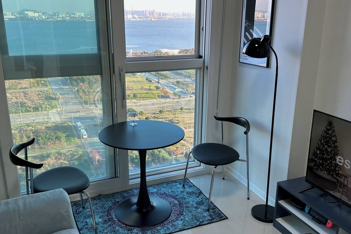 A modern living room with a large window offering a city view, a round table with two chairs, a floor lamp, and a TV stand with electronic devices.