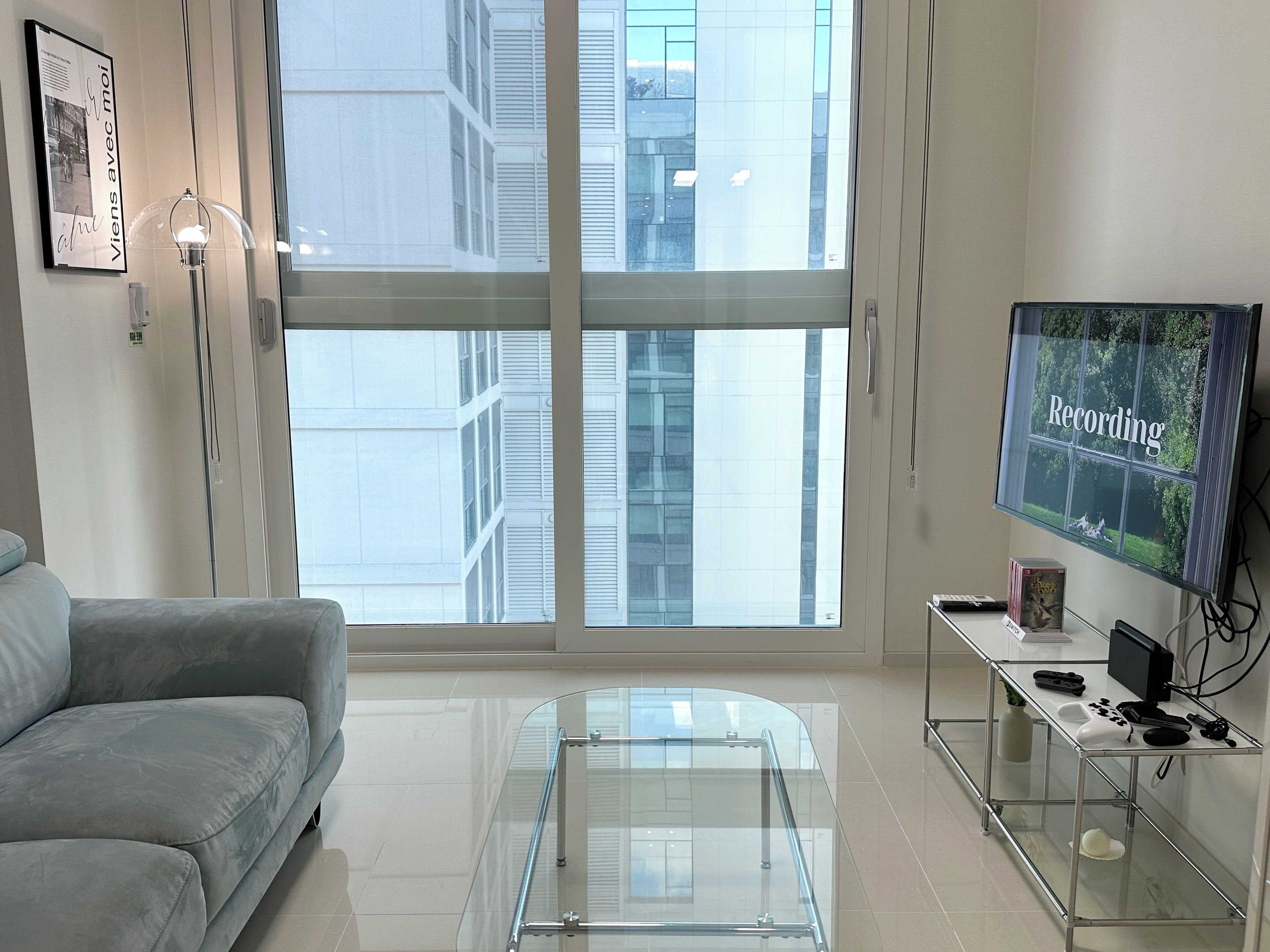 A modern living room with a grey sofa, a glass coffee table, a TV on a stand, and a large window with a city view.