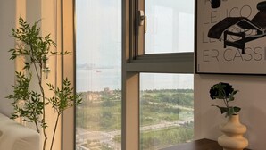 Een moderne kamer met een groot raam, een houten bureau, een witte stoel, een potplant en een kunstwerk in een lijst aan de muur.