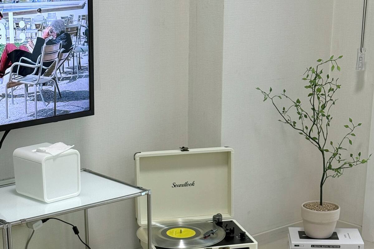 A modern room with a glass table, a soundproofed music player, a potted plant, and a television displaying a scene with people.