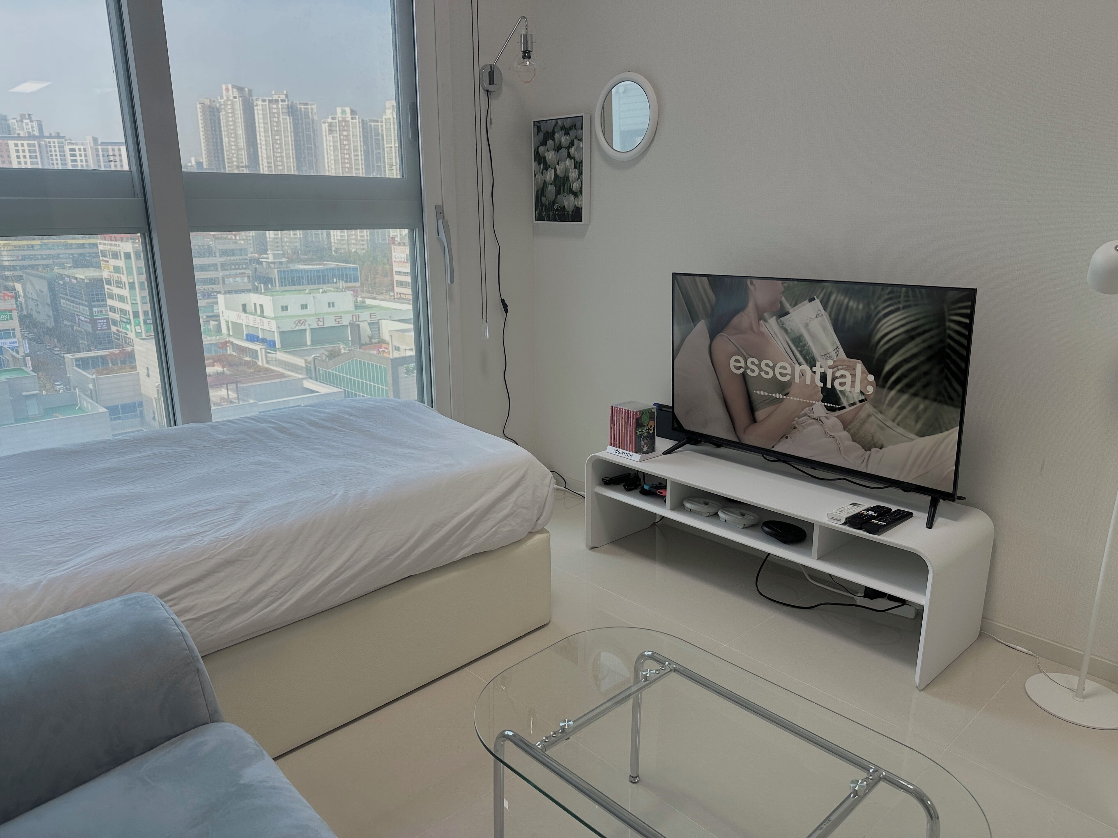 A modern hotel room with a large bed, a TV on a white stand, a glass coffee table, and a city view through the window.