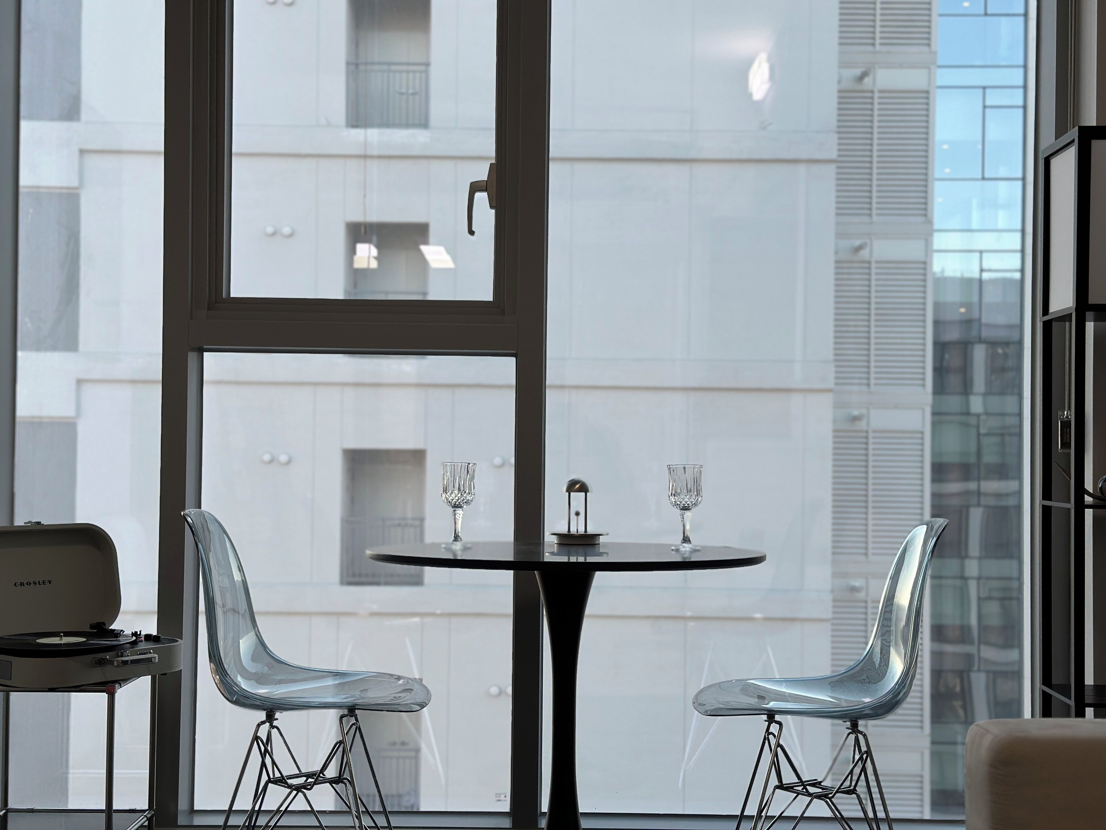 A modern interior with a glass table and two transparent chairs, a turntable, and a view of a white building through large windows.