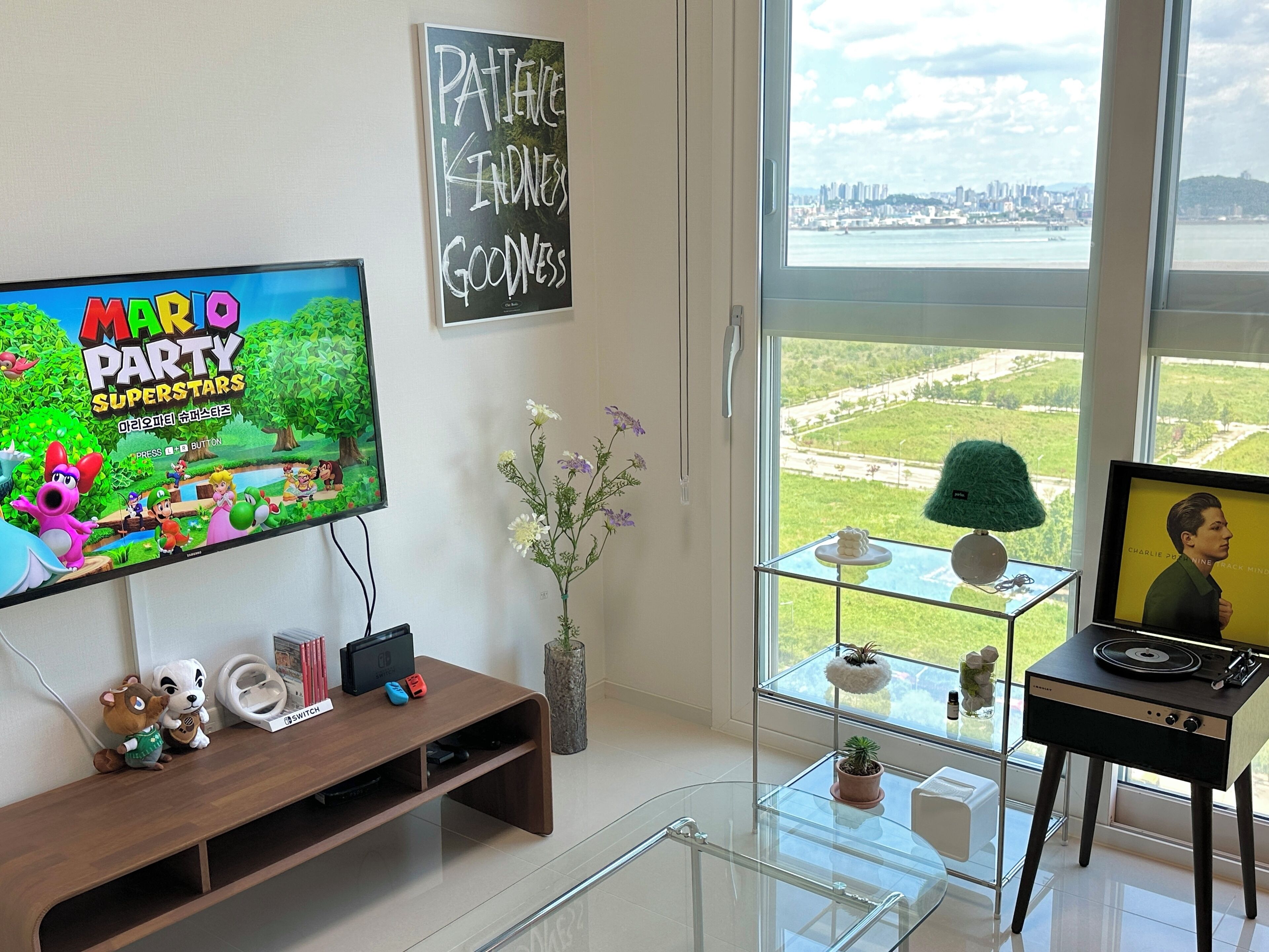 Sala de estar con una televisión mostrando 'Mario Party Superstars', un mueble de madera para equipos electrónicos, una mesa de centro de vidrio y vistas a un paisaje urbano a través de ventanas amplias.