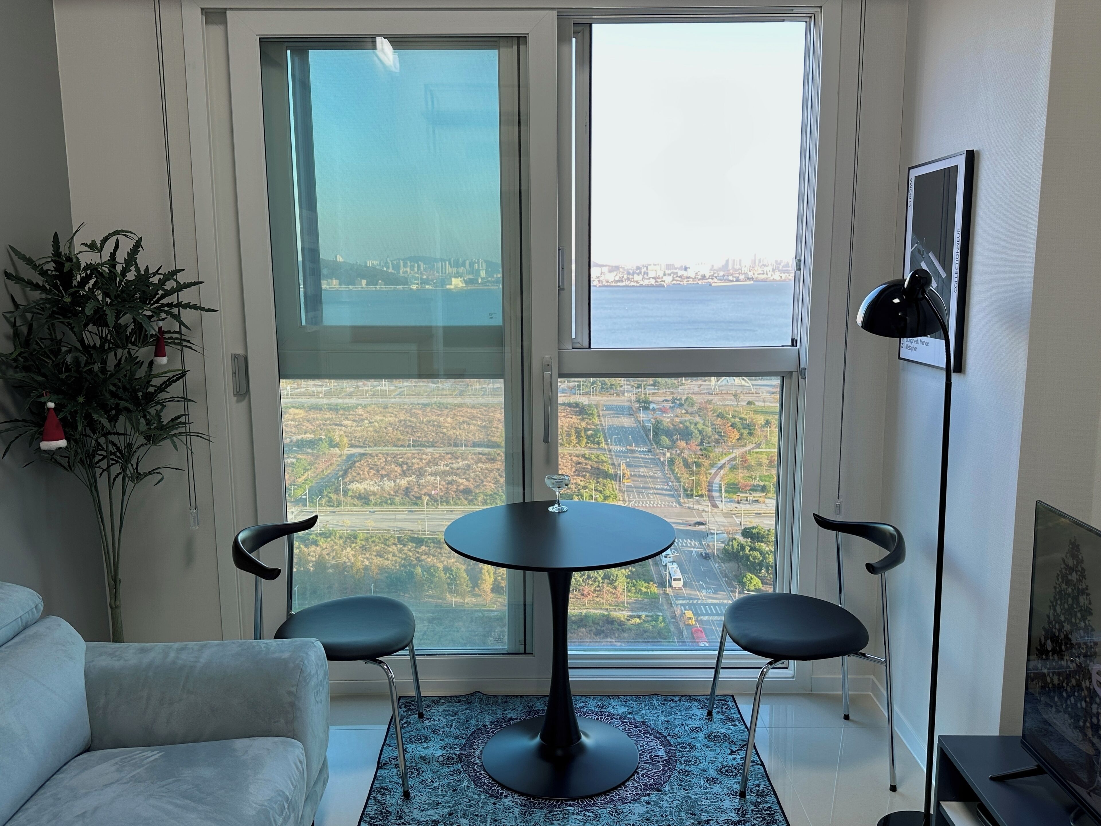 A modern living room with a large window offering a city view, a round table with two chairs, a sofa, a floor lamp, and a potted plant.