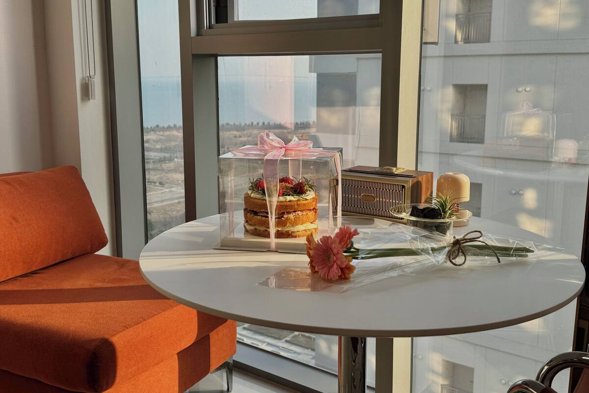 A room with a round table, a clear glass display case with a cake, a wooden radio, and a potted plant.