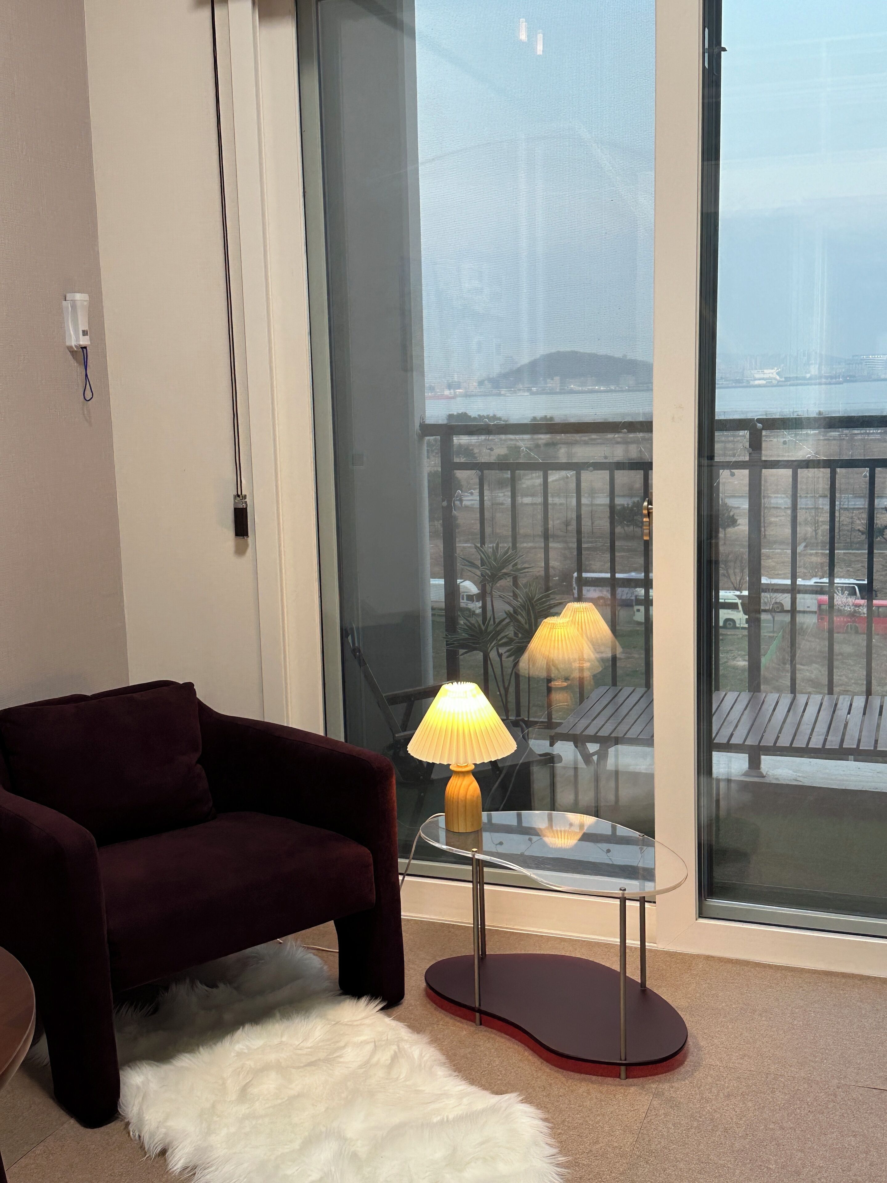 A living room with a dark-colored sofa, a glass table with a lamp, and a view of a balcony with a railing and a body of water in the background.