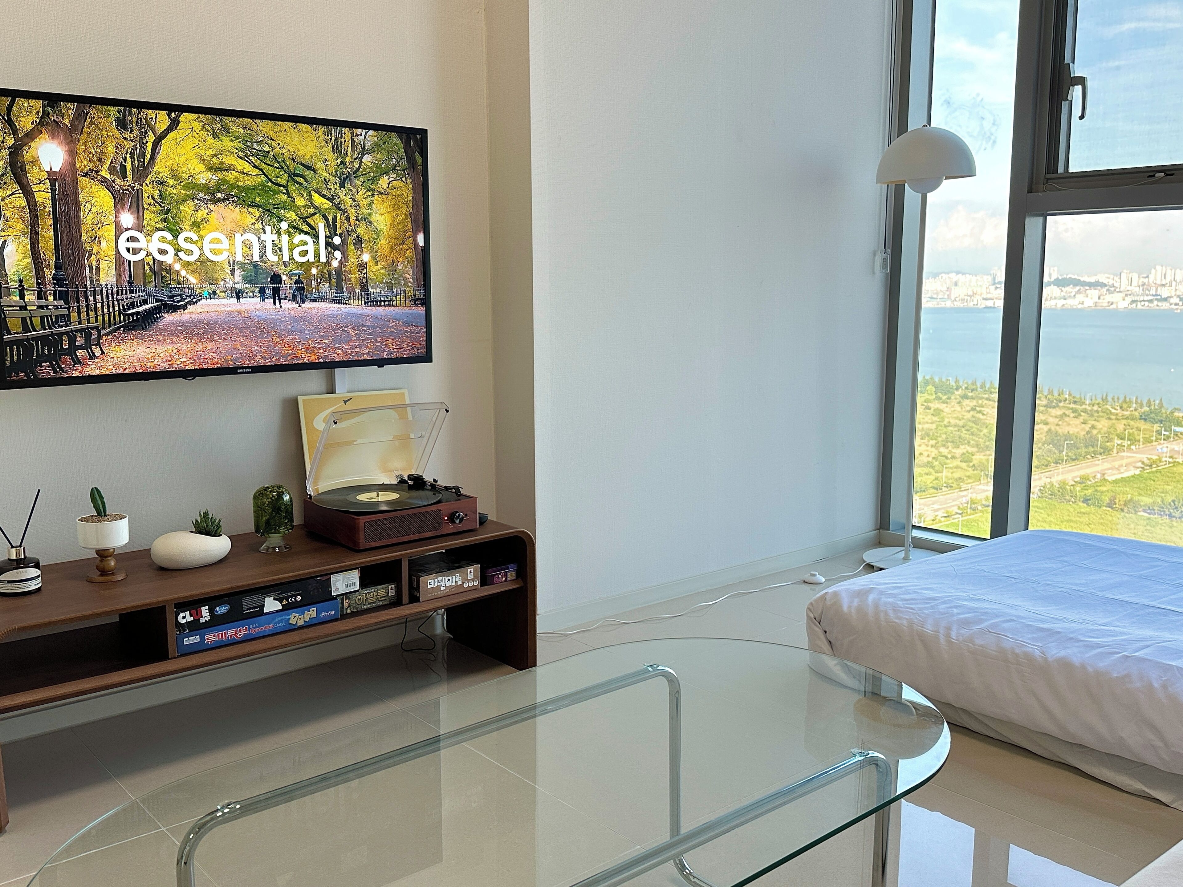 A modern hotel room with a large TV mounted on the wall, a glass table, a bed, and a view of a cityscape through large windows.