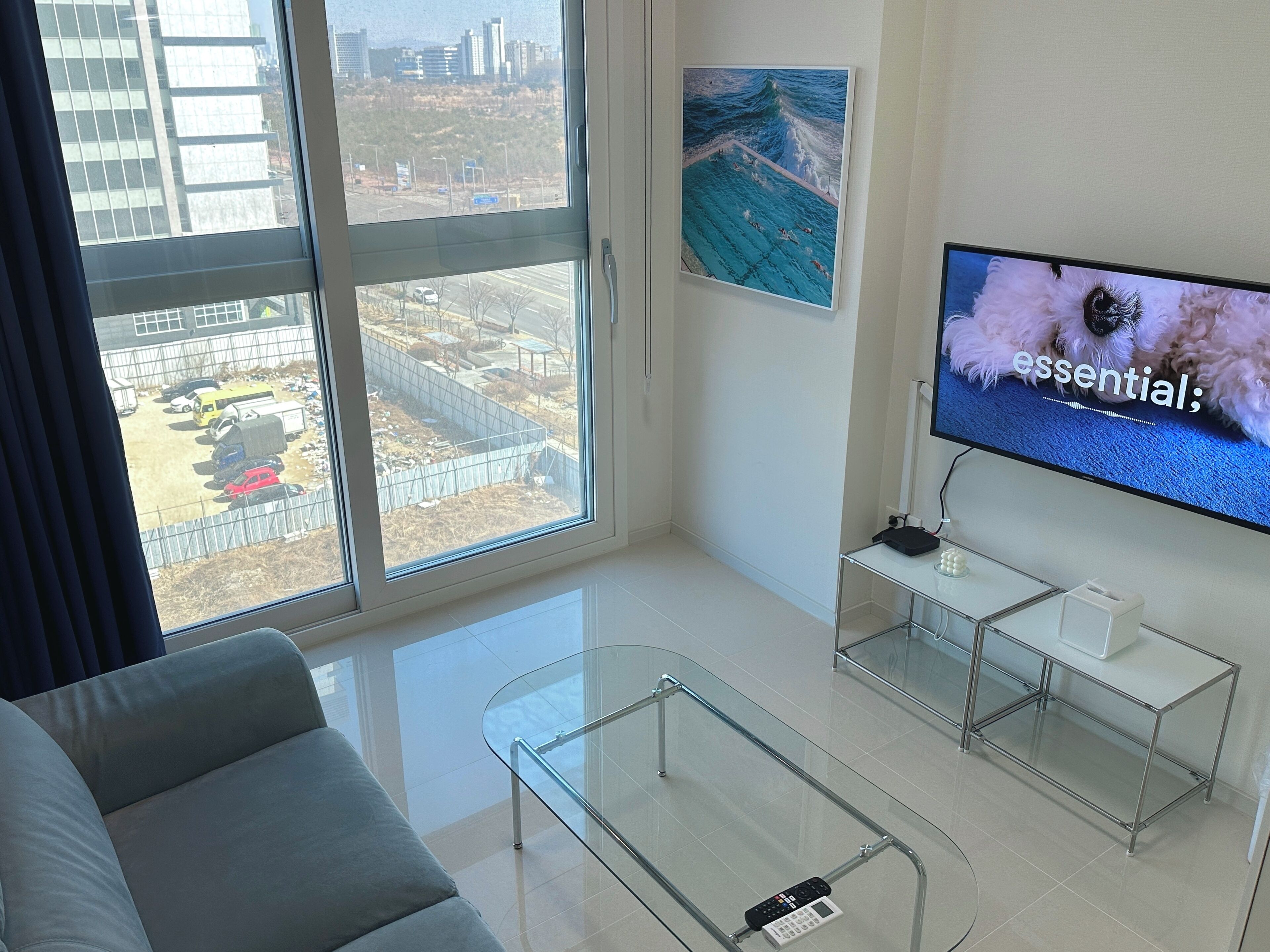 A modern living room with a large window, a sofa, a glass coffee table, and a TV displaying the word "essential".